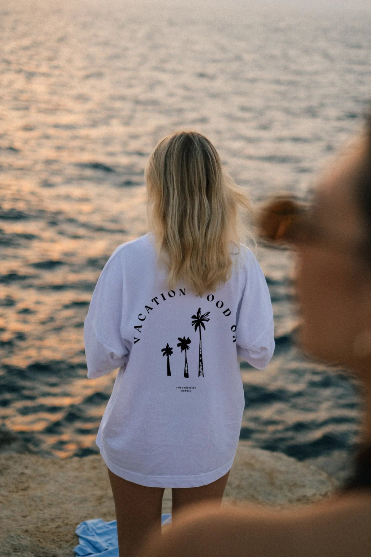 Frau mit weißem Vacation Mood On T-Shirt steht bei Sonnenuntergang auf Felsen