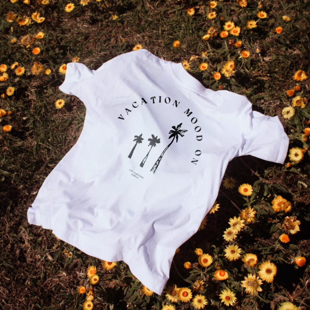 Weißes T-Shirt mit „Vacation Mood On“-Print liegt dekorativ auf einer sommerlichen Blumenwiese