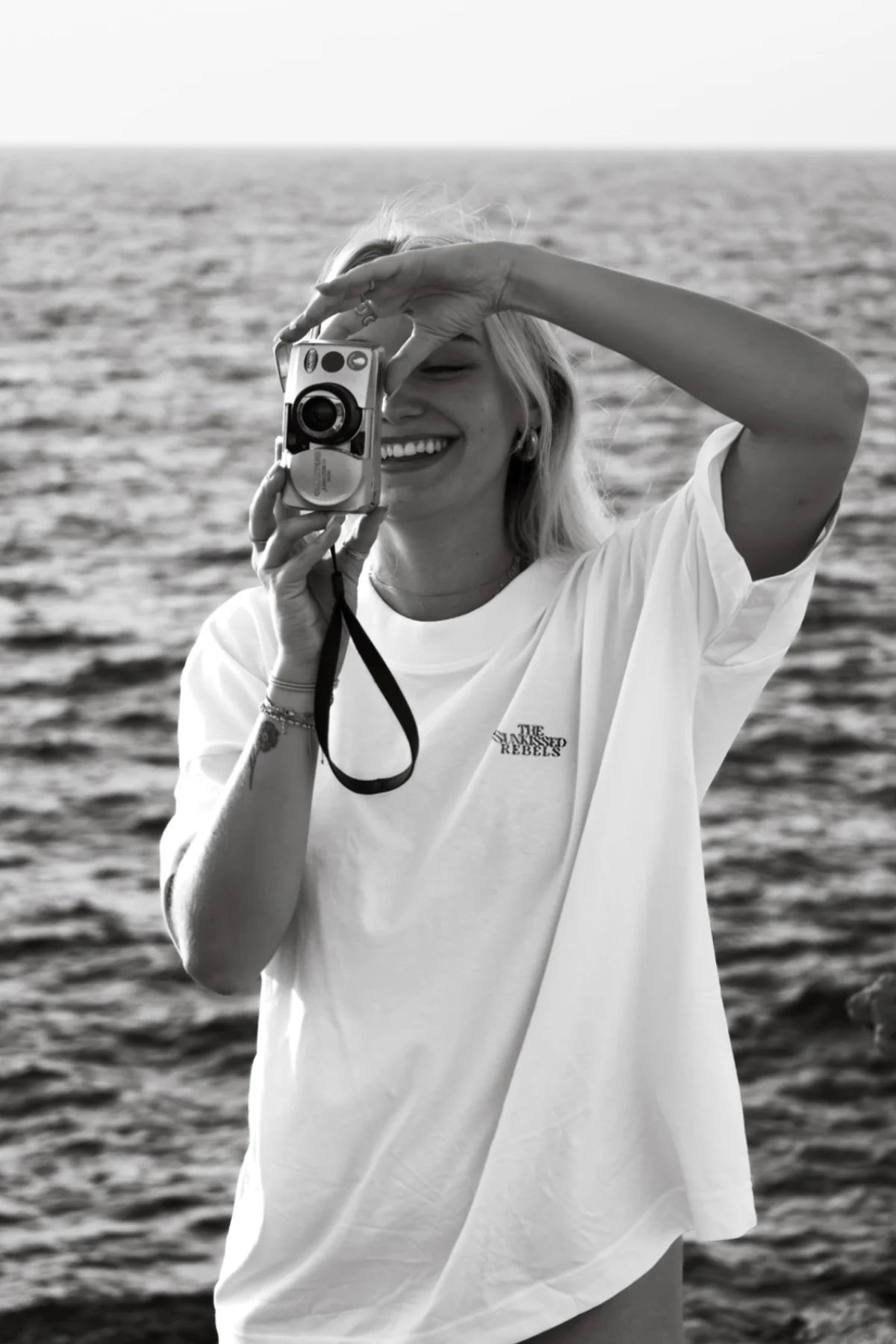 Frau in weißem Oversized T-Shirt hält Kamera am Meer – Momentaufnahme