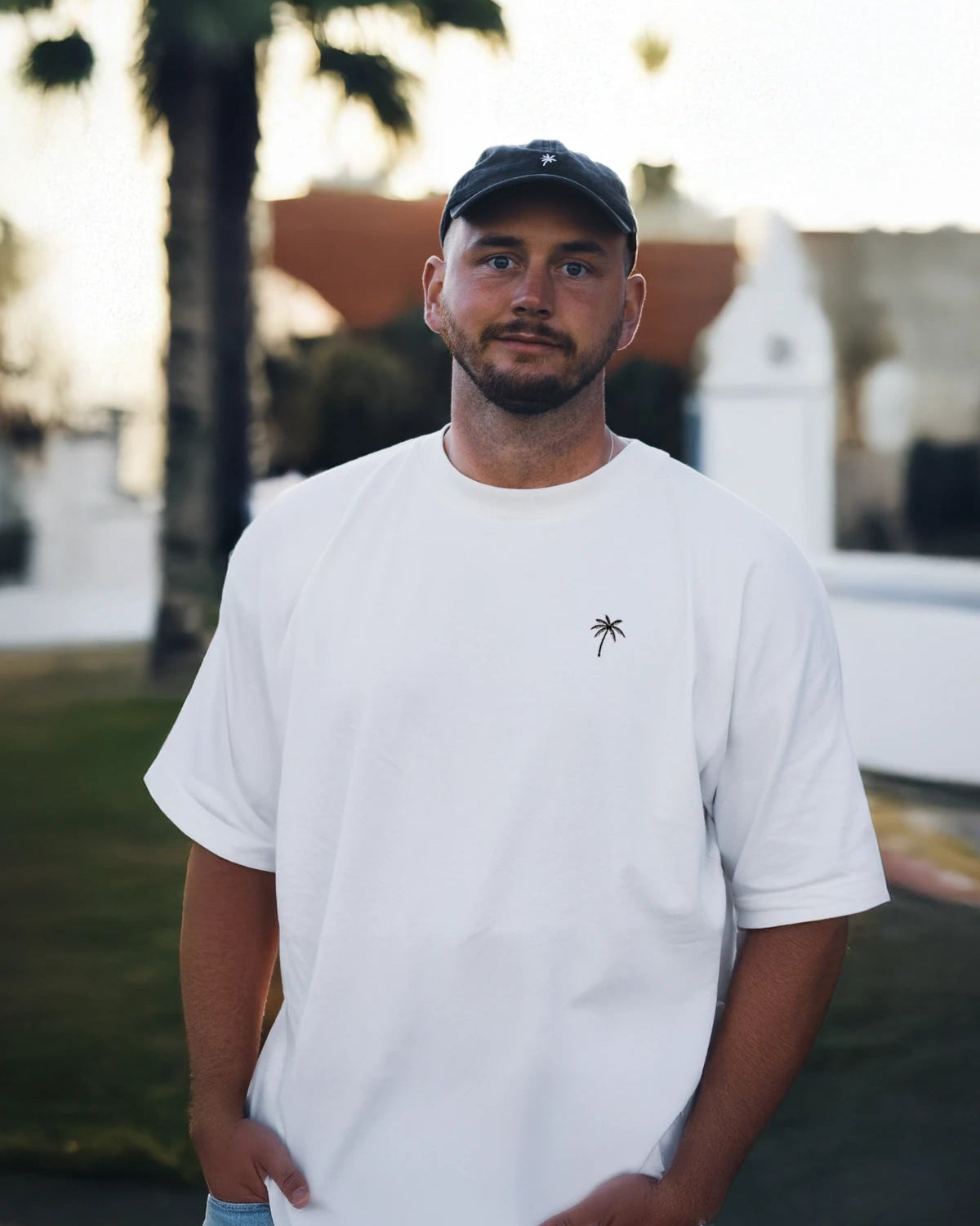 Mann mit weißem Oversized T-Shirt von The Sunkissed Rebels und Palmen-Stick – lässiger Urban Look.