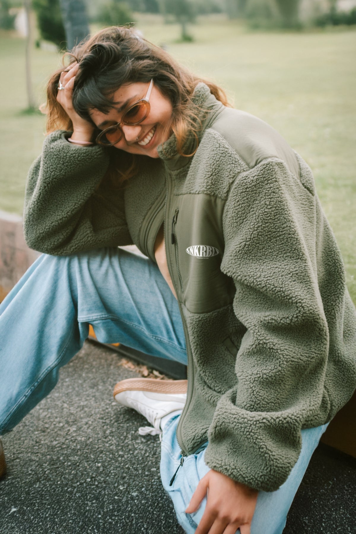 Frau sitzt auf Bordstein und lacht, trägt khaki / grüne fleece jacke