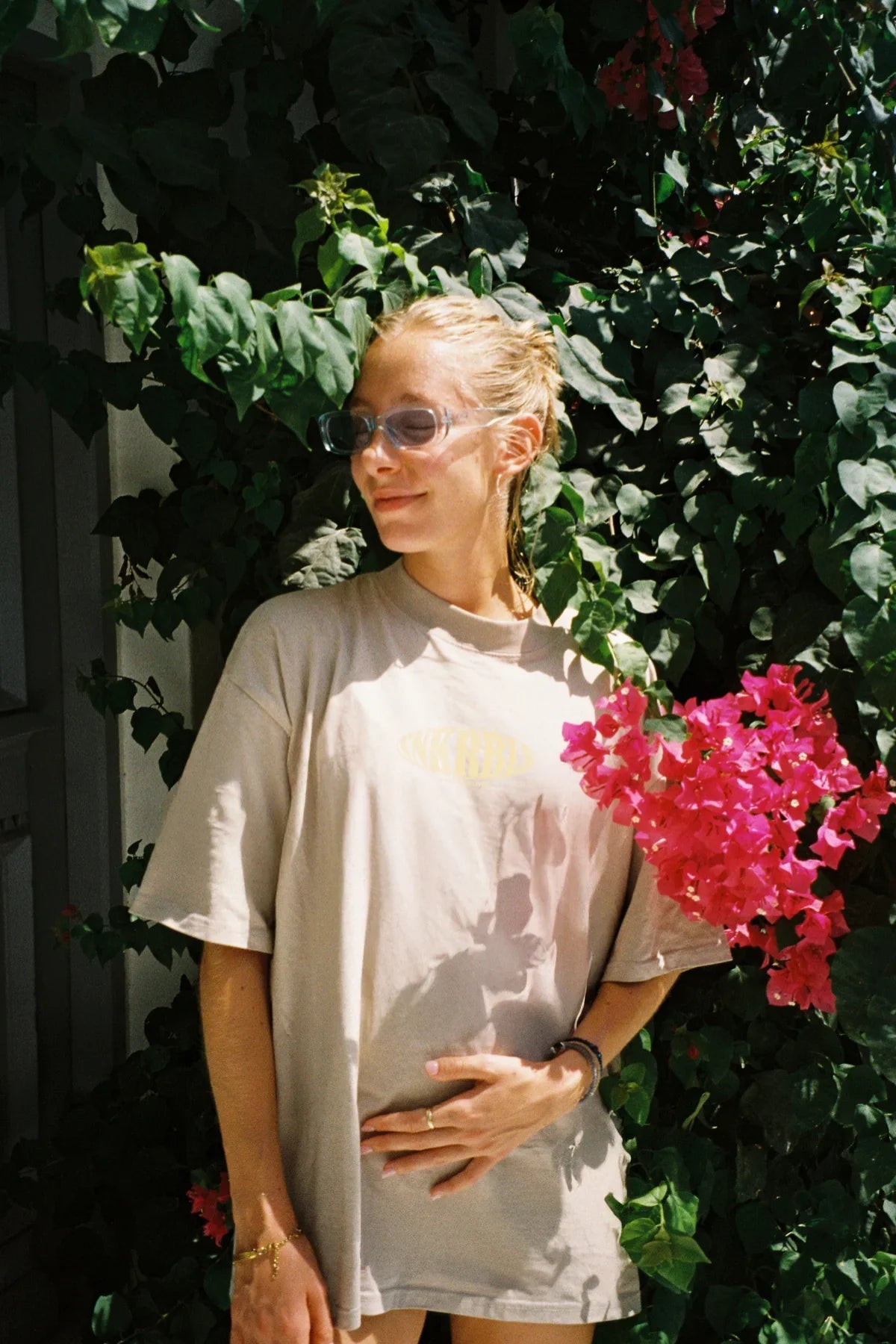 Frau im grauen Oversized T-Shirt steht vor Blumenwand bei Sonennschein