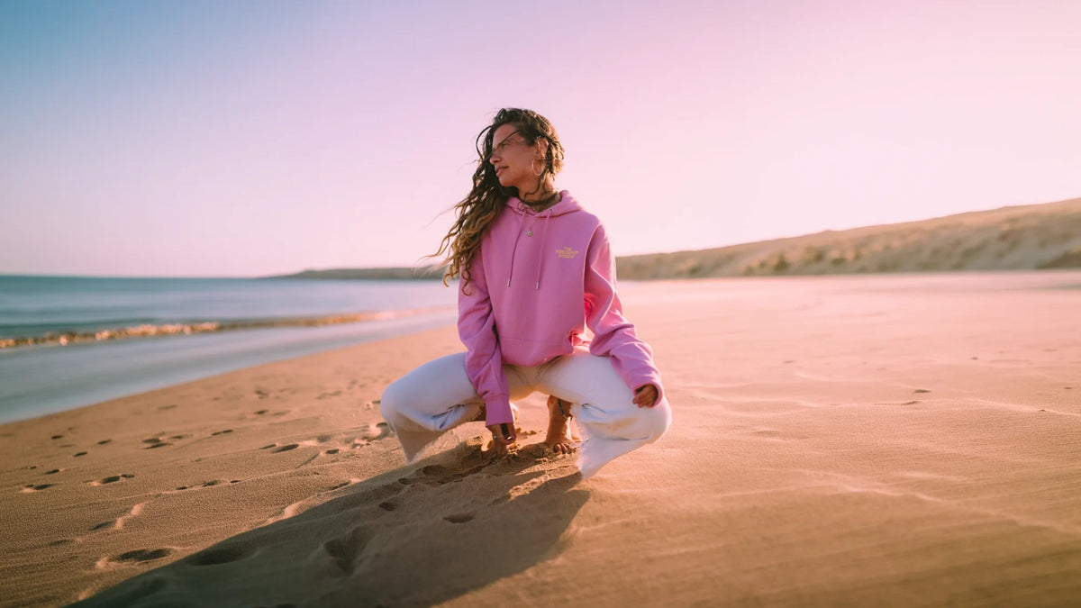 Frau im pinken The Sunkissed Rebels Hoodie am Strand bei Sonnenuntergang – nachhaltige Vacation Wear im Surf Lifestyle.