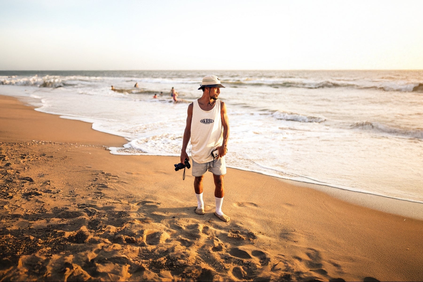 Mann am Strand mit Kamera in der Hand trägt Tank top von The Sunkissed Rebels