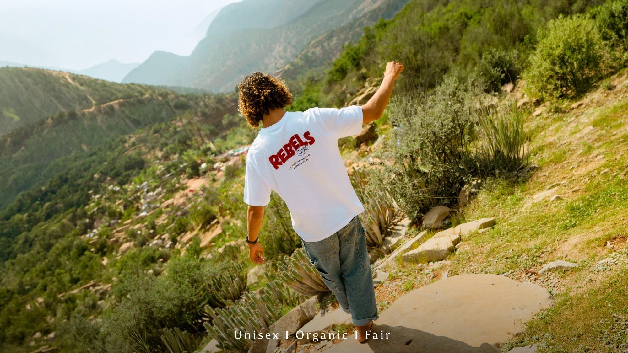 Person mit weißem REBELS Organic Unisex T-Shirt auf einem Bergpfad in natürlicher Outdoor-Landschaft