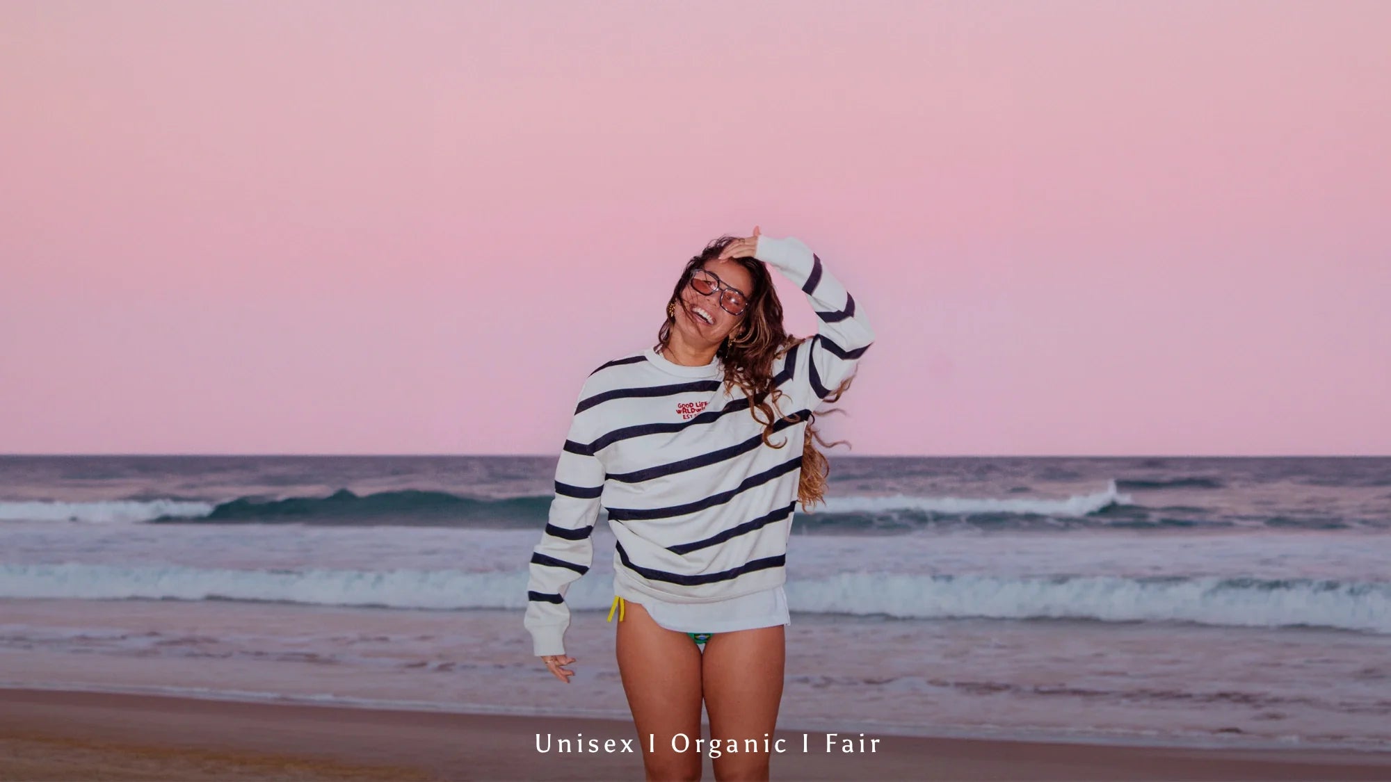 Weibliches Model trägt gestreiften Organic Sweater am Strand bei pinkem Sonnenuntergang und entspannter Surf Atmosphäre.
