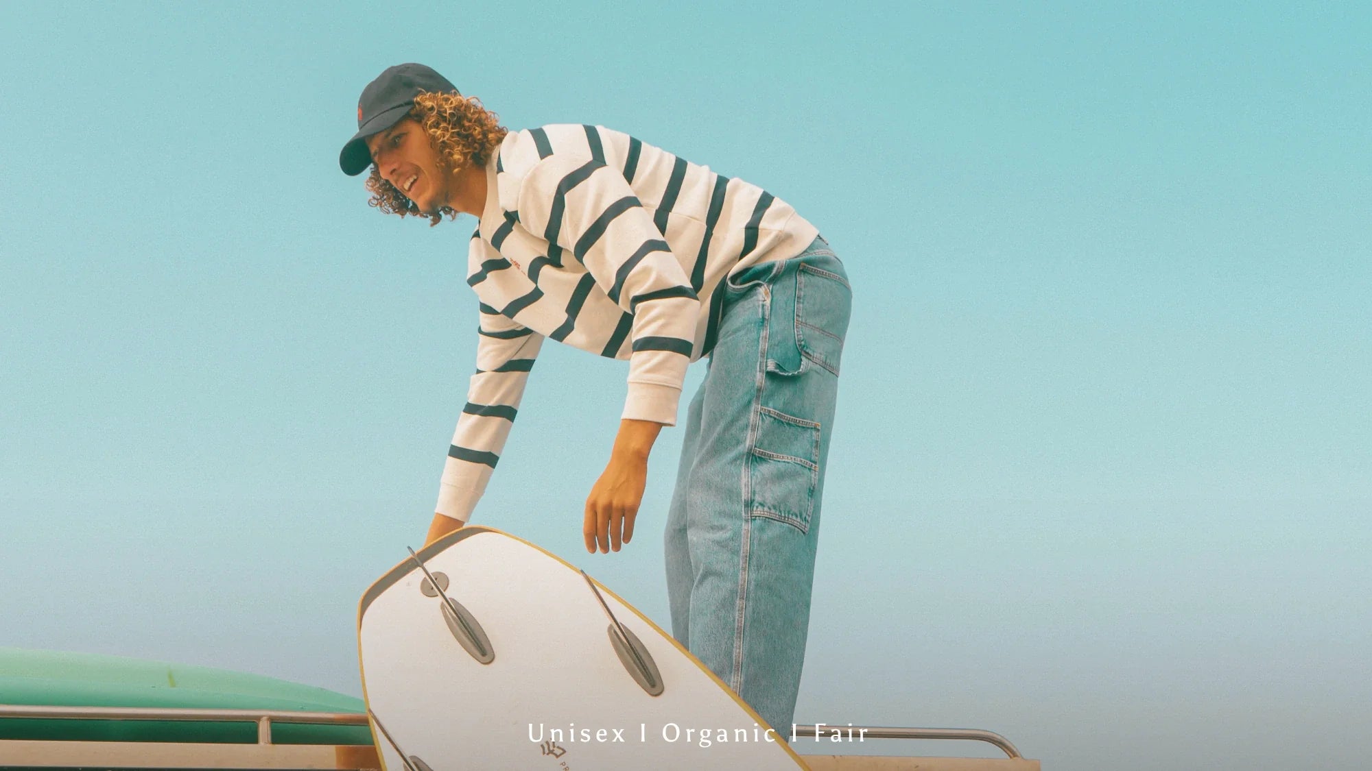 Männliches Model mit gestreiftem Organic Longsleeve und Jeans auf Surfboard bei sonnigem Beach Lifestyle Shooting.