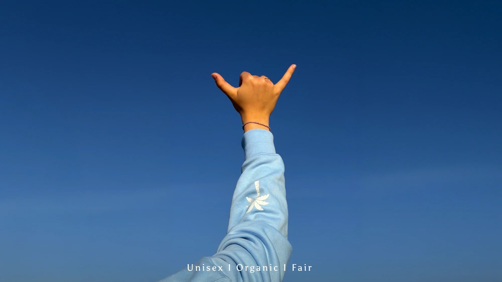 Shaka-Handzeichen mit hellblauem Organic Fairwear Hoodie vor tiefblauem Himmel