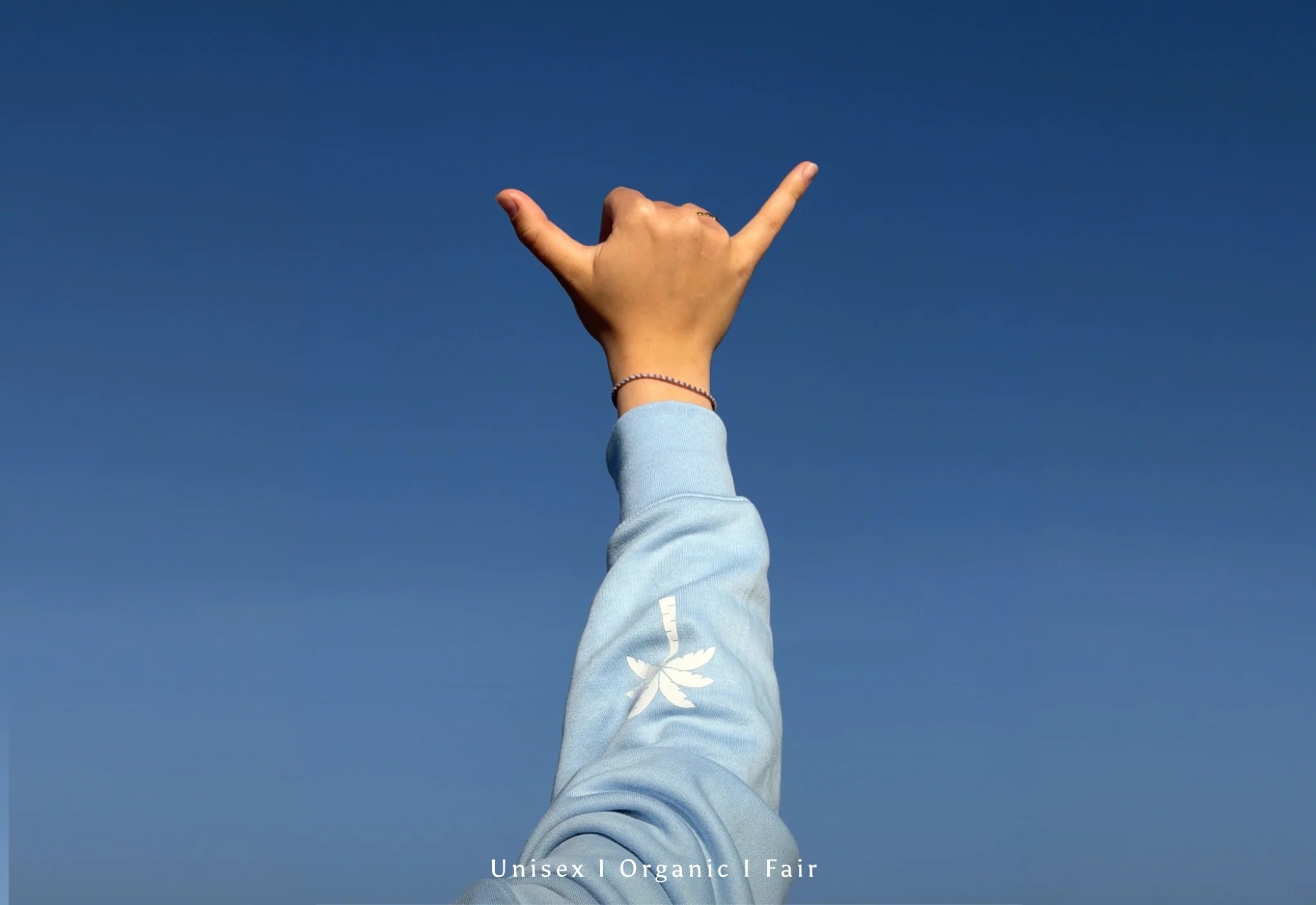 Hand zeigt Shaka Zeichen vor klarem blauen Himmel, minimalistisches Lifestyle Bild mit hellblauem Ärmel, Unisex Organic Fair Fashion Ästhetik