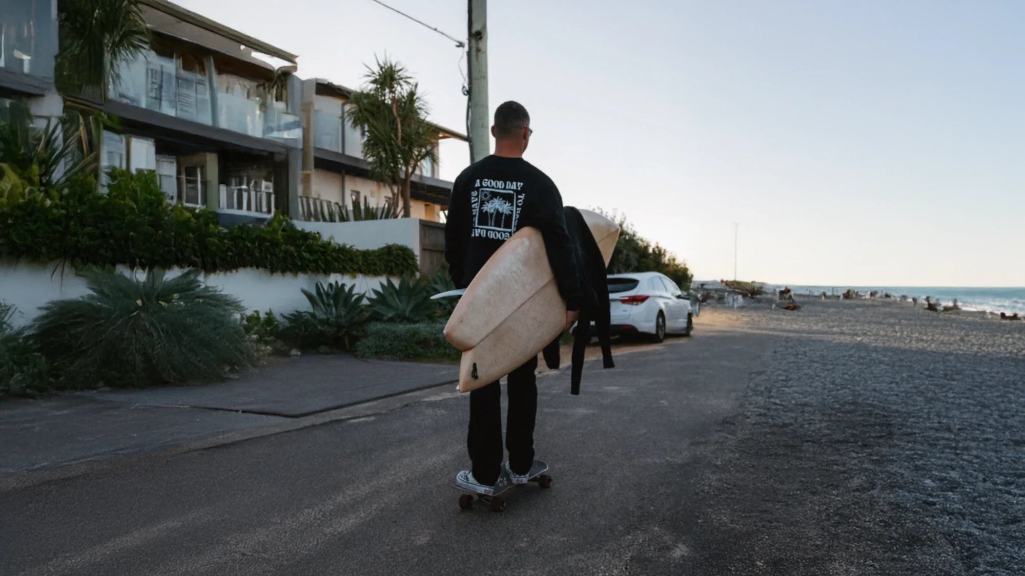 Mann fährt auf einem Skateboard mit Surfboard unterm Arm am Strand entlang – lässiger Streetwear-Look von The Sunkissed Rebels.