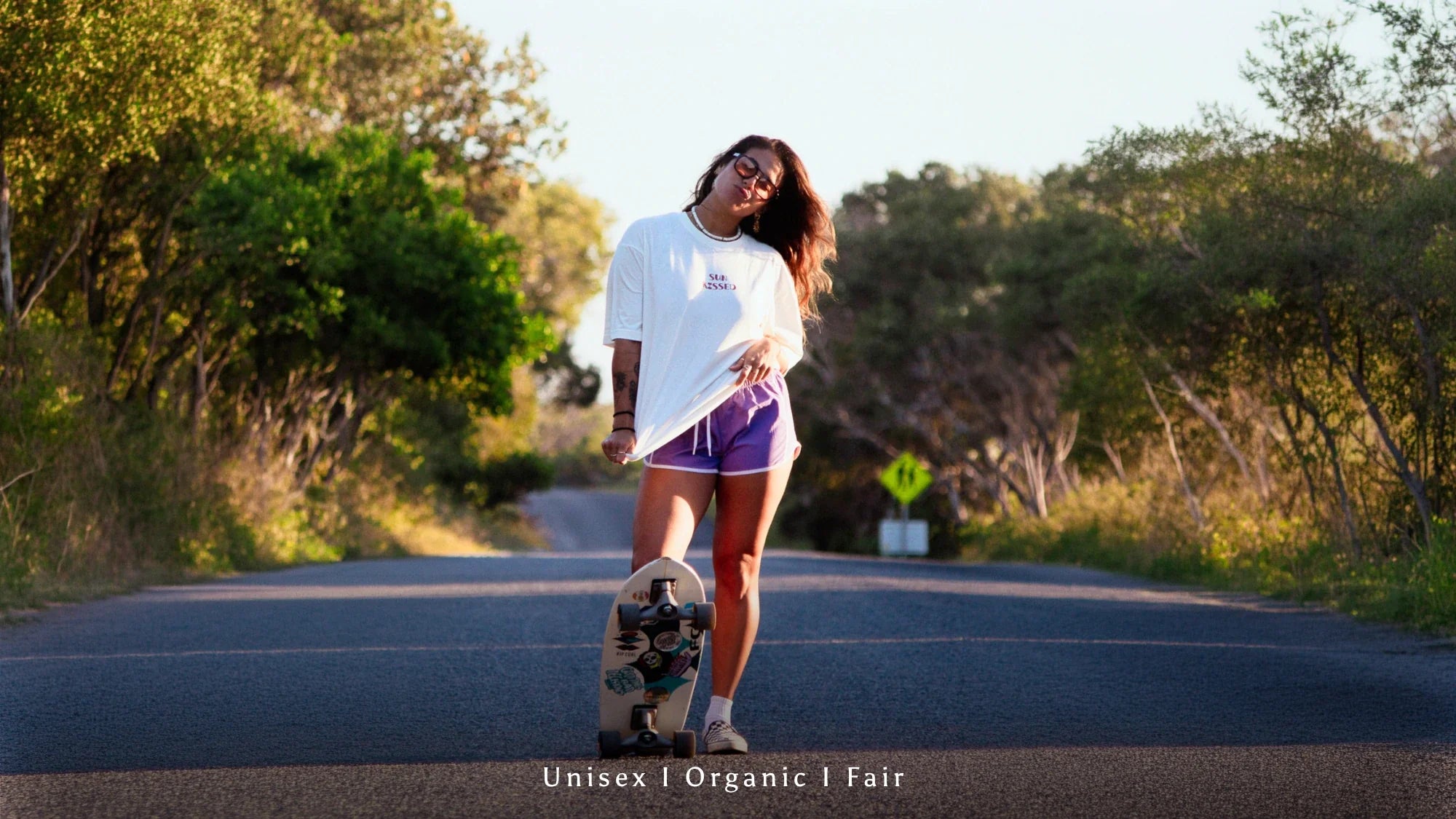 Junge Frau mit Skateboard auf einer Straße trägt ein weißes Unisex Organic Shirt von Sunkissed Rebels bei Sonnenuntergang im Skater Lifestyle Look. 