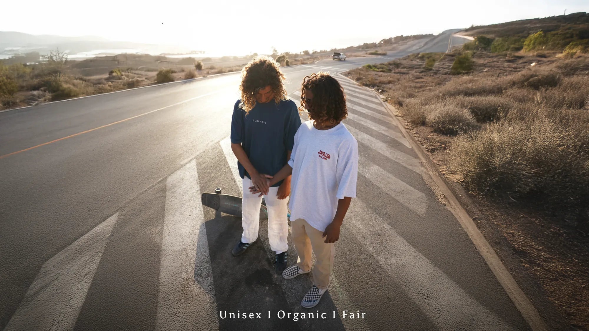Zwei junge Models von The Sunkissed Rebels mit lockigen Haaren stehen auf einer sonnigen Landstraße, tragen lässige Unisex-Organic Shirts und verkörpern einen natürlichen, fairen und authentischen Lifestyle.