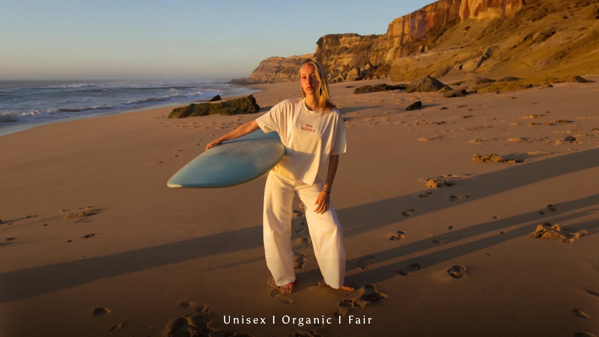 Surferin mit Surfboard in weißer Organic Unisex Kleidung am Strand während Golden Hour bei Sonnenuntergang