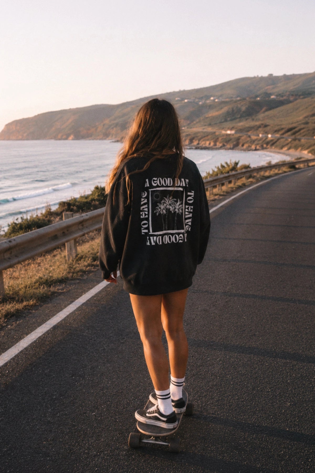 Frau trägt den schwarzen A Good Day Sweater mit großem Rückenprint beim Longboarden auf einer sonnigen Küstenstraße am Meer.