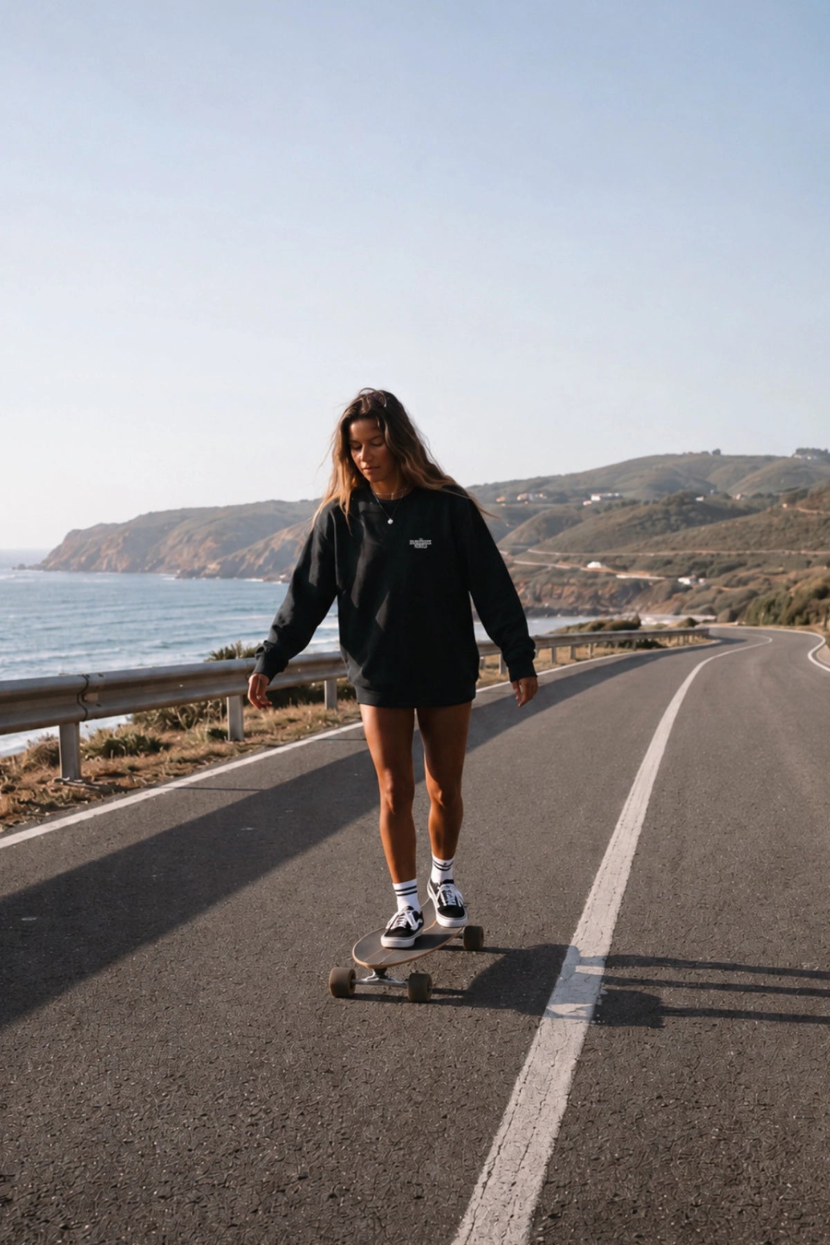Frau trägt den schwarzen A Good Day Sweater beim Longboarden auf einer Küstenstraße mit Meerblick.