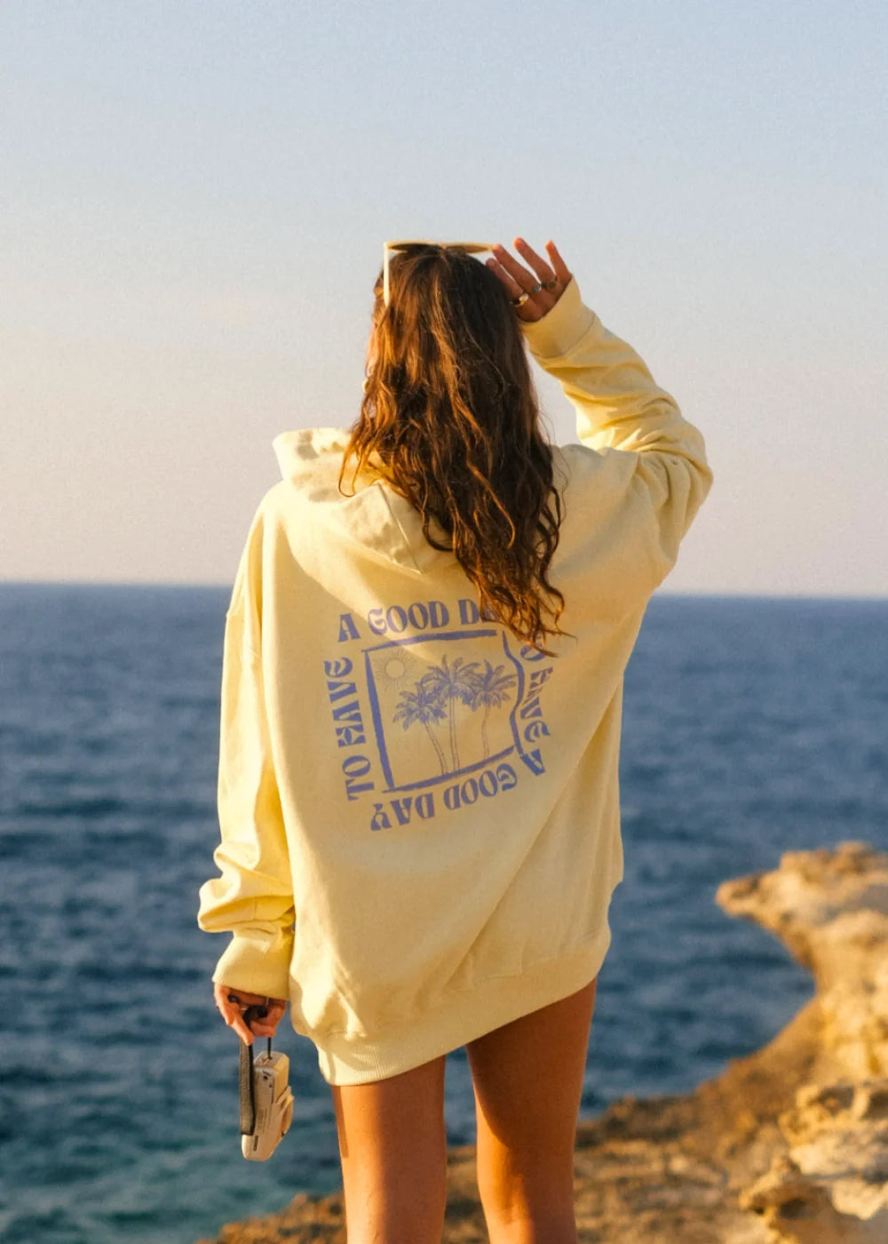Rückansicht einer Frau im A Good Day Hoodie in VButter Yellow von The Sunkissed Rebels mit großem Rückenprint an einer