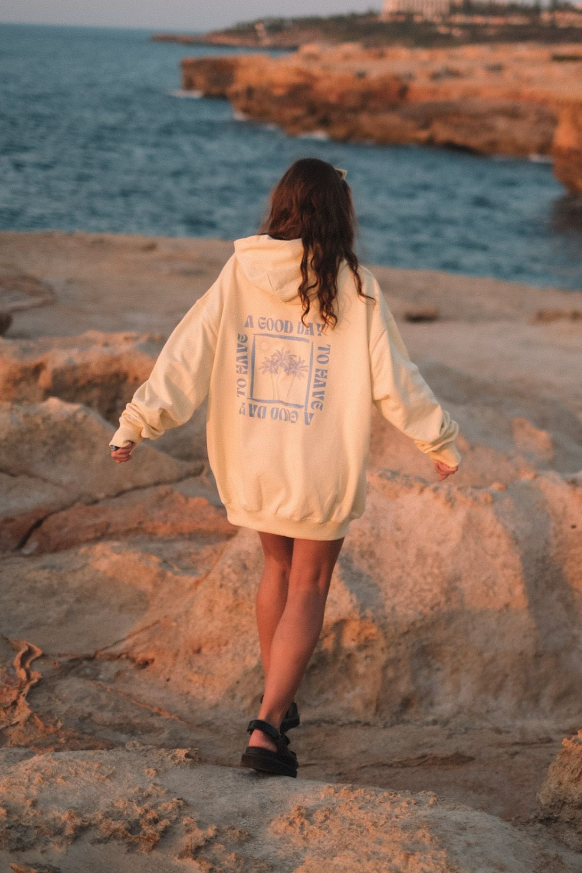 Frau trägt A Good Day Oversized Sweater von The Sunkissed Rebels am Strand im Sonnenuntergang. 