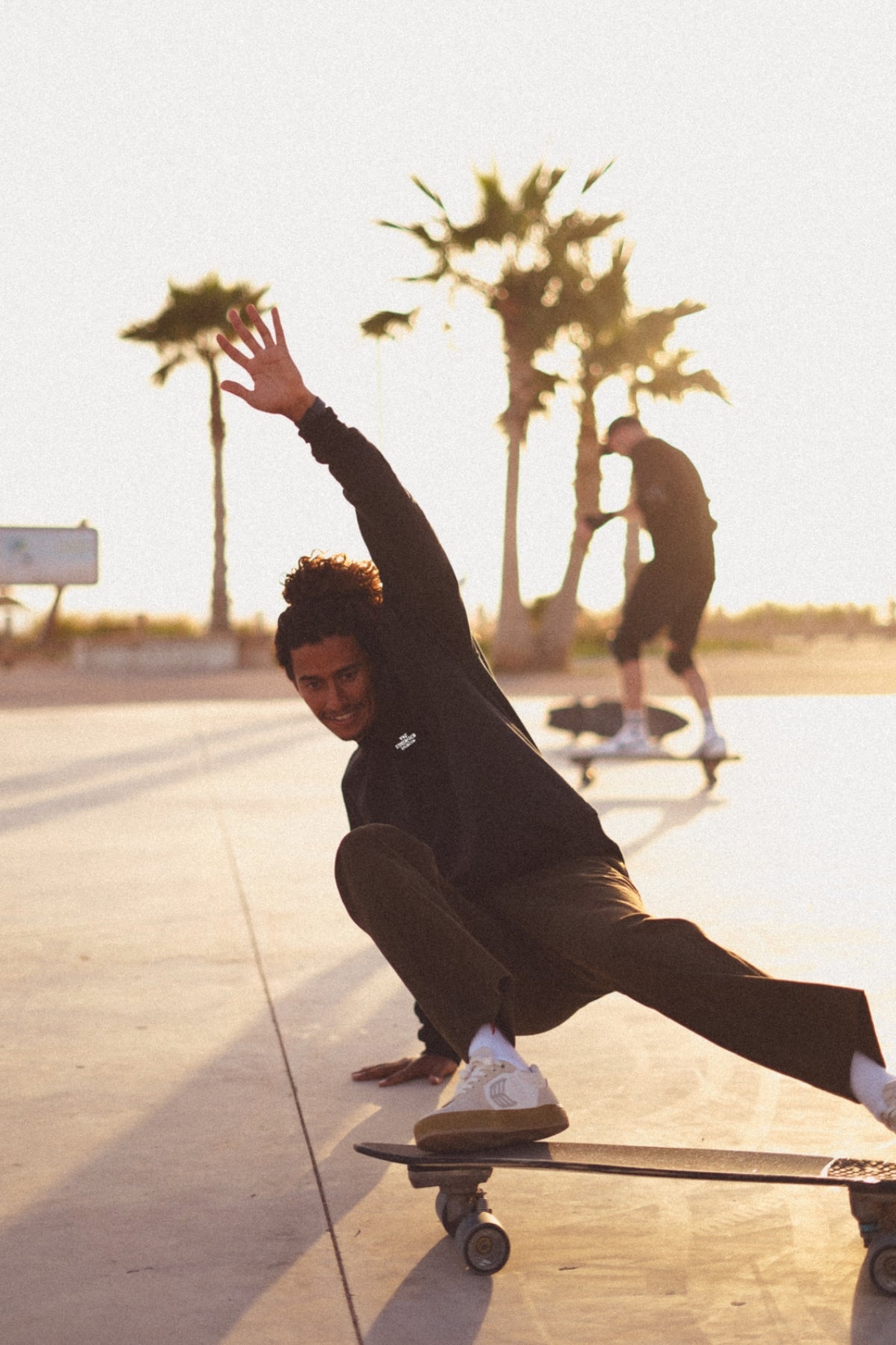 Man fährt Longboard am Strand, im Hintergrund sind Palmen zu sehen. Er trägt einen schwarzen Oversized Sweater von The Sunkissed Rebels. 