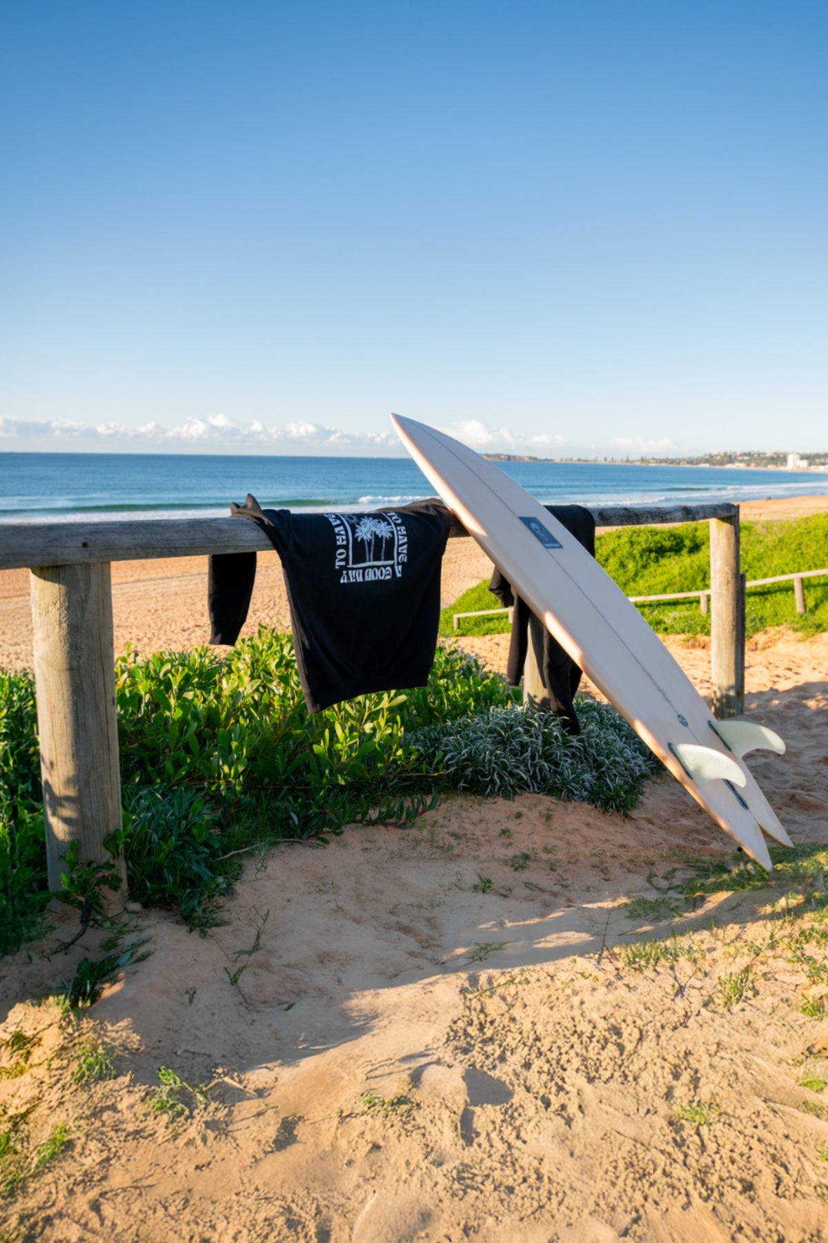 Der Sweater A Good Day von The Sunkissed Rebels hängt über einen Zaun am Strand neben einem Surfbrett. 