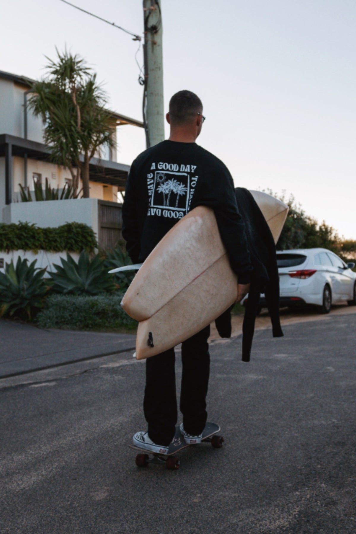 Skateboarder trägt einen schwarzen Oversized Sweater und hat ein Surfbrett unterm Arm geklemmt. 