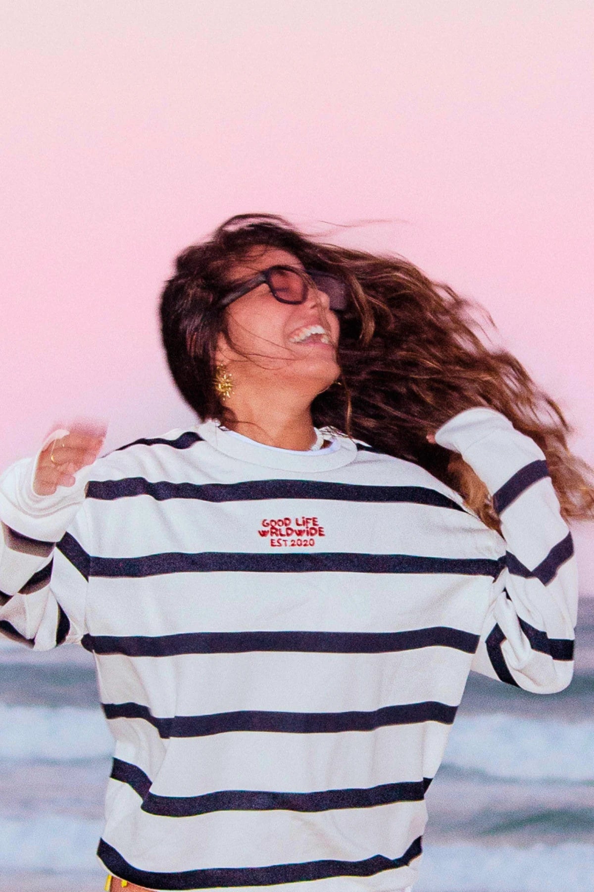 Gestreifter Dockside Rebel Sweater Cream mit Good Life Worldwide Stickerei bei einem Fotoshooting am Strand.