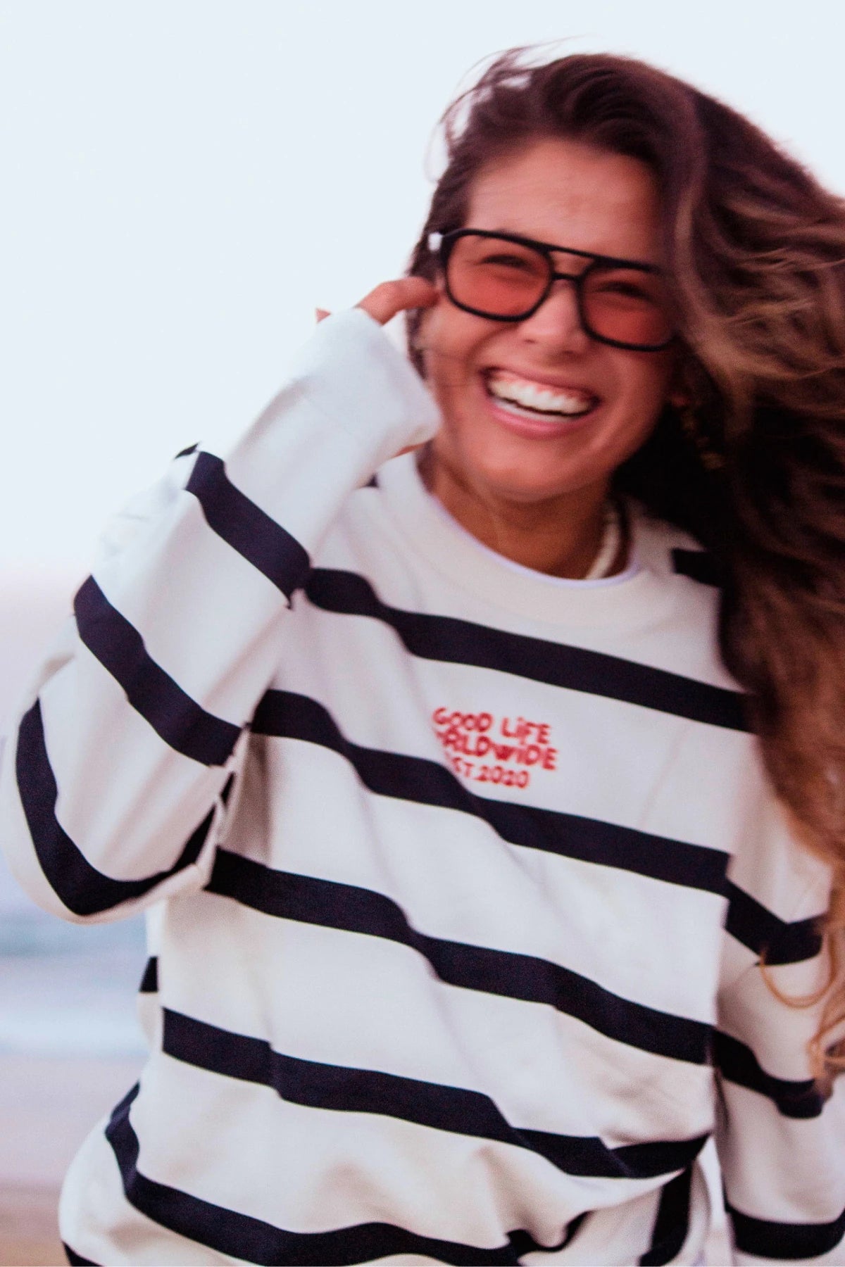 Frau trägt den Dockside Rebel Sweater Cream am Strand bei Sonnenuntergang mit maritimen Streifen und entspannter Ausstrahlung. 