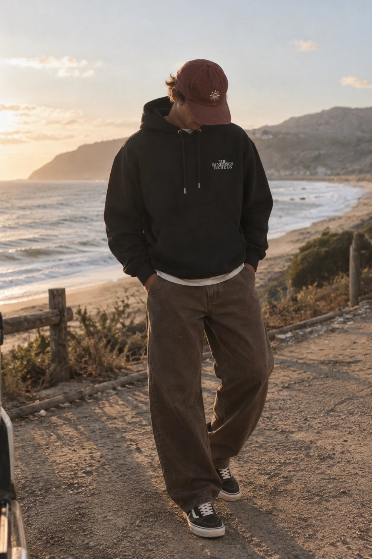 Frontansicht eines Mannes in einem schwarzen Kapuzenpullover der Marke „The Sunkissed Rebels“. Er lehnt lässig an einem weissen Van, im Hintergrund das Meer bei Sonnenuntergang.