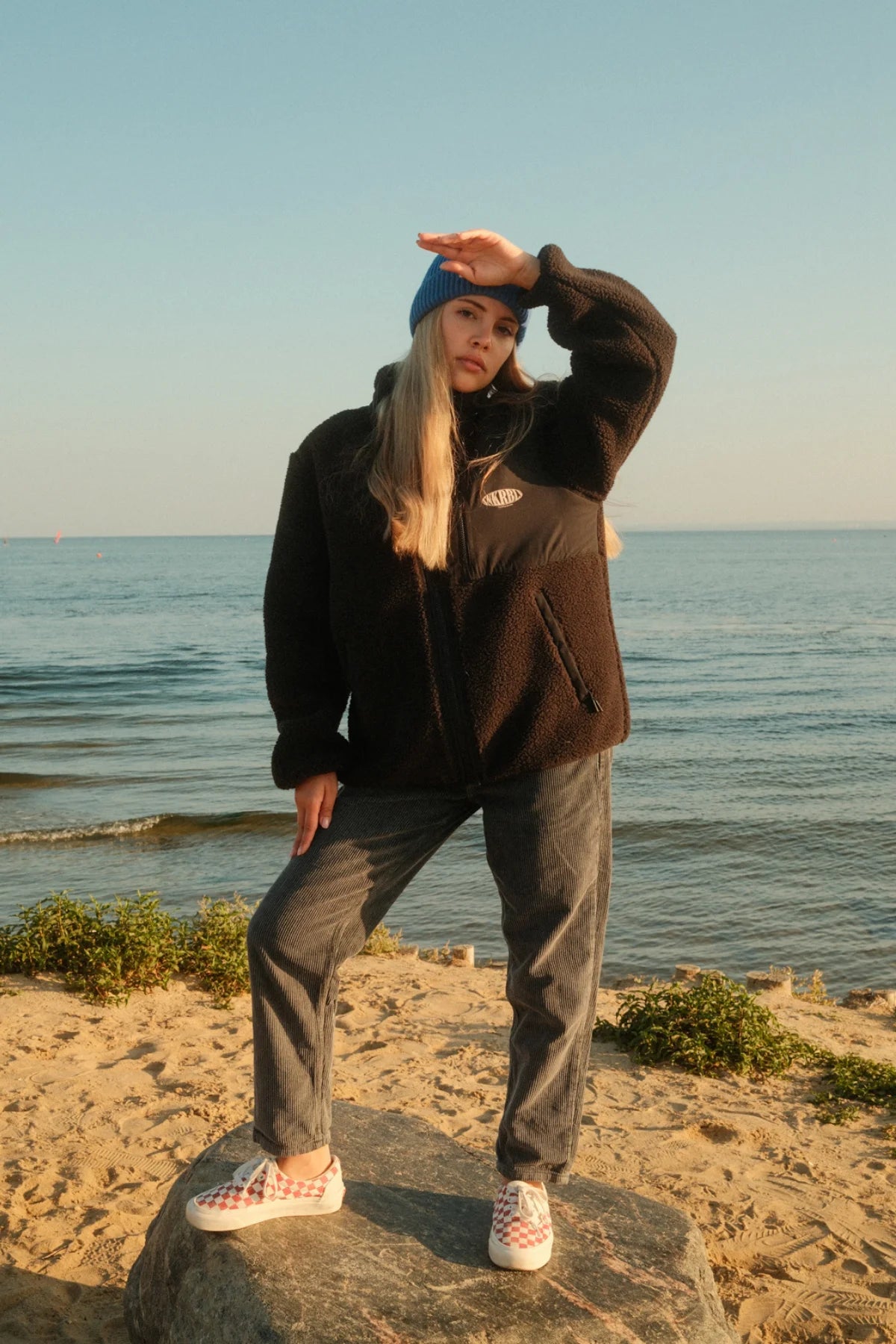 Schwarze Teddy Fleecejacke von The Sunkissed Rebels kombiniert mit Kordhose und blauer Beanie am Meer.