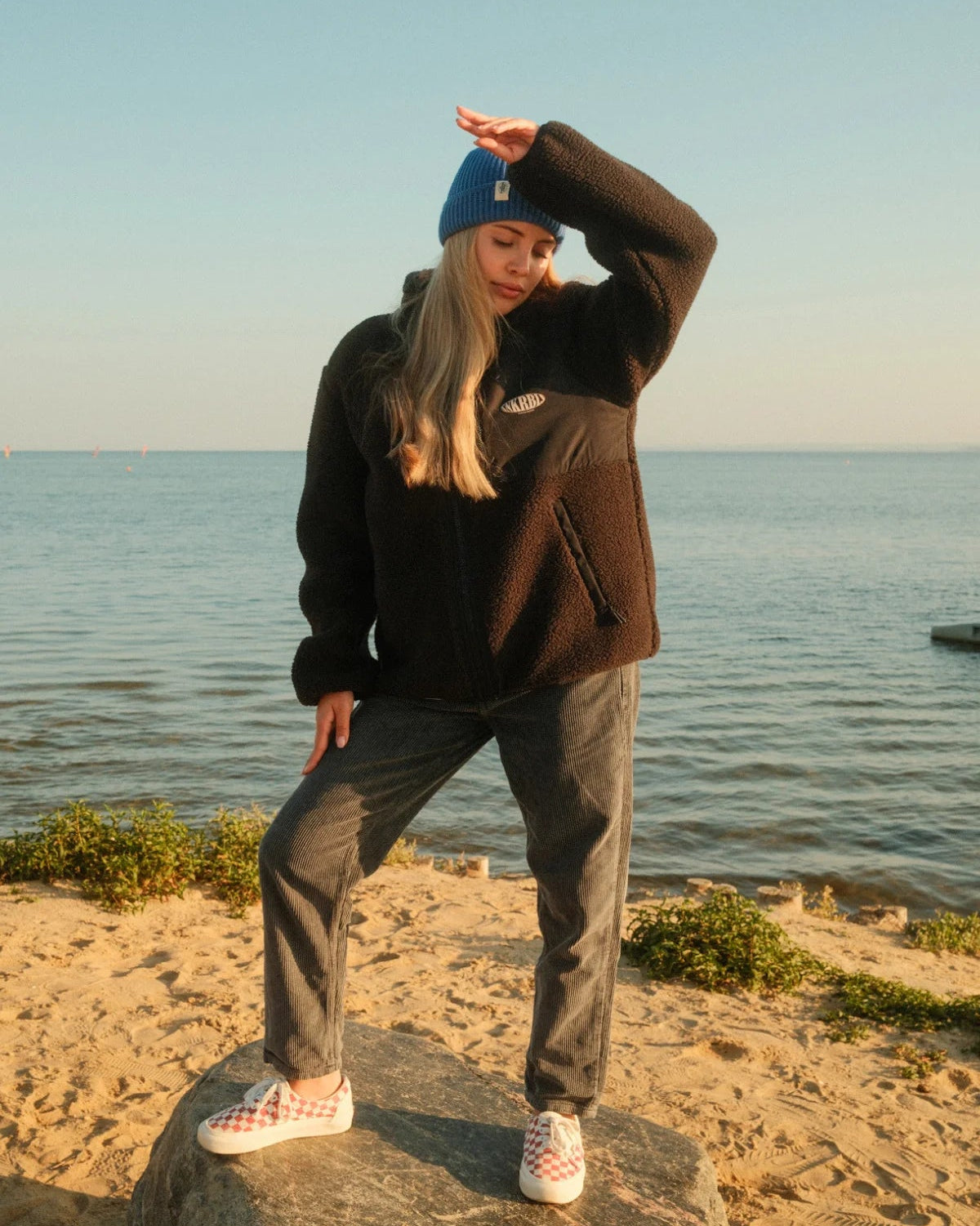 Frau am Strand mit schwarzer Fleece Jacket von The Sunkissed Rebels, blauer Beanie und SNKRBL Brust-Logo.