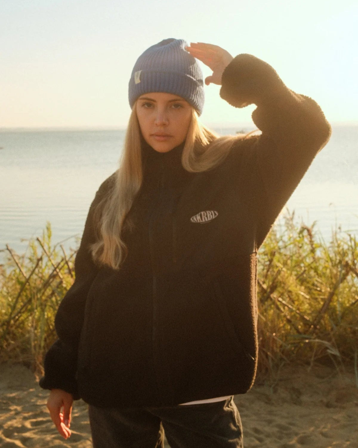 Entspannter Coastal Look mit geschlossener schwarzer Fleecejacke von The Sunkissed Rebels im Gegenlicht.