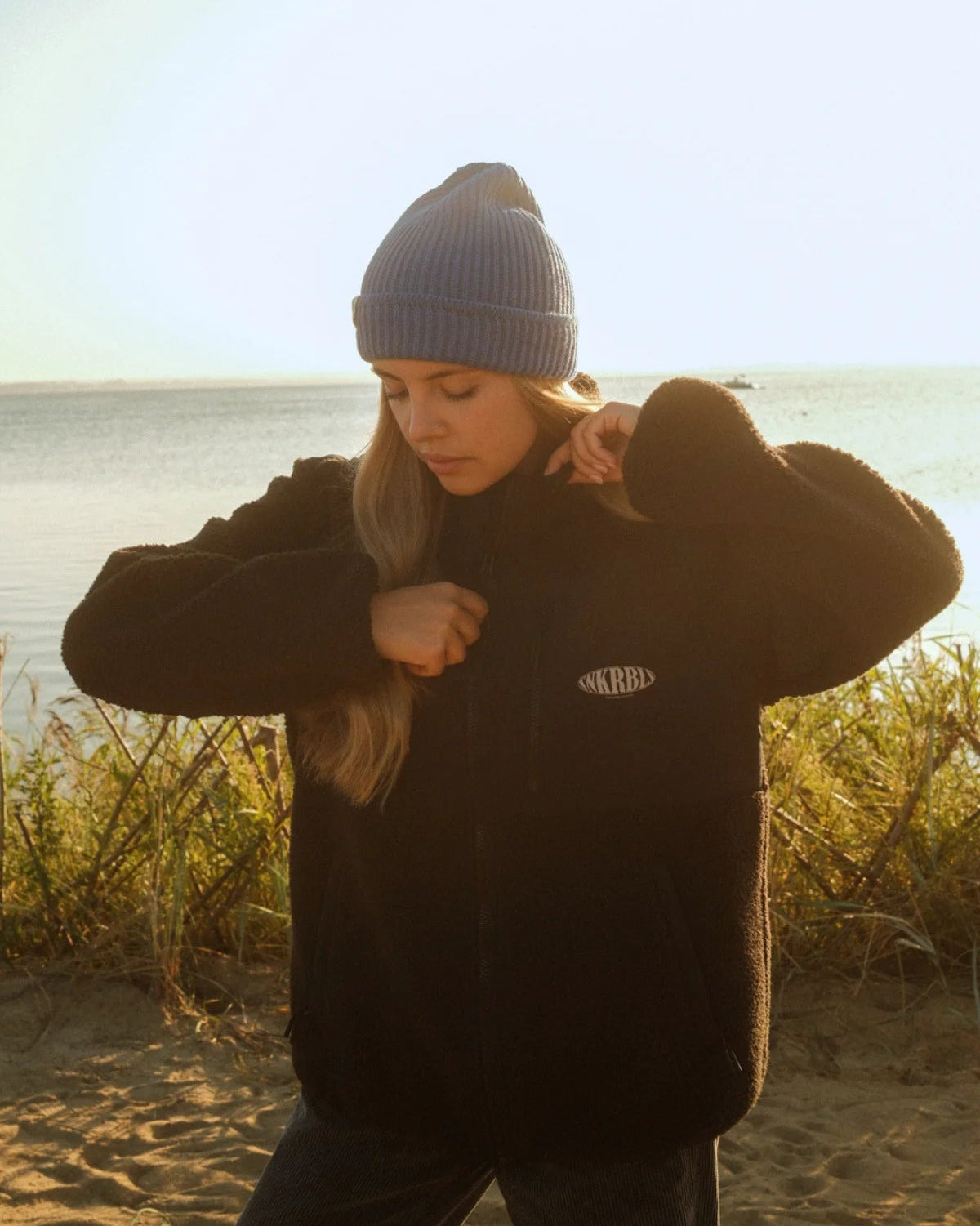 Frau schließt die schwarze Fleece Jacket von The Sunkissed Rebels mit sichtbarem SNKRBL Logo-Print am Strand.