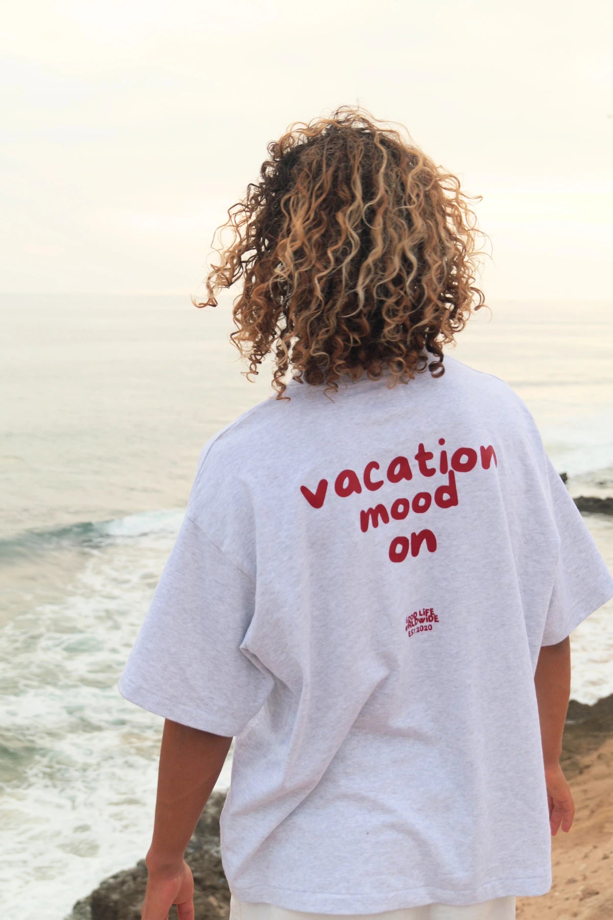 Detailaufnahme des Goodlife Heavy T-Shirts in Cool Grey mit großem roten Vacation Mood On Rückenprint an der Küste.