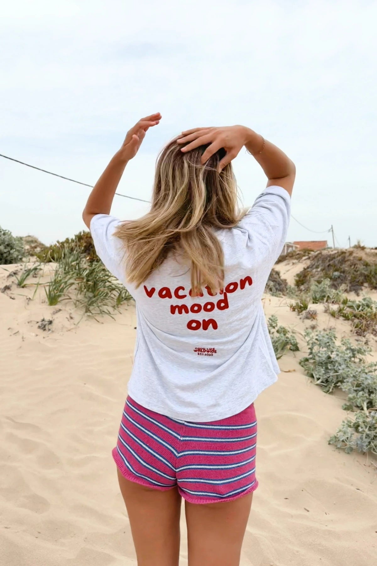Rückenansicht des Goodlife Heavy T-Shirts in Cool Grey mit rotem Vacation Mood On Print in den Sanddünen.