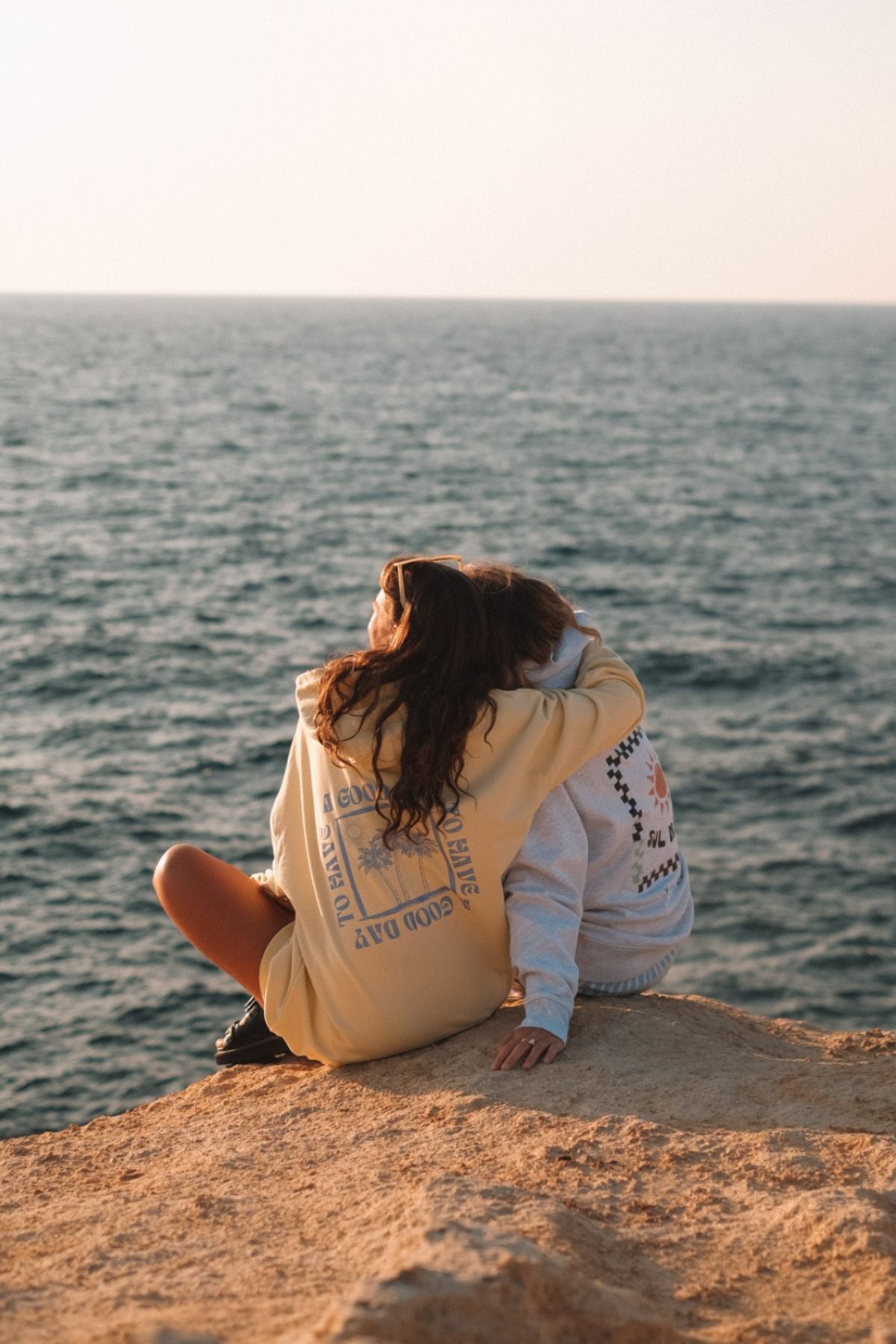 Zwei Frauen sitzen am Strand, halten sich in den Armen und schauen den Sonnenuntergang. Beide tragen Hoodies von The Sunkissed Rebels.