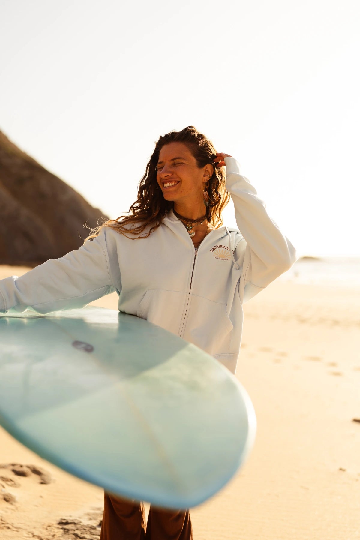 Lächelnde Surferin trägt den Iconic Women Zip-Hoodie Pacific Chill am Sandstrand mit Surfboard bei Sonnenuntergang.