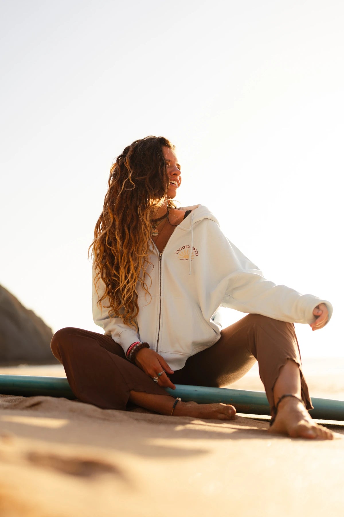 Frau sitzt entspannt am Strand auf einem Surfboard und trägt den Iconic Women Zip-Hoodie Pacific Chill im Beach-Look.