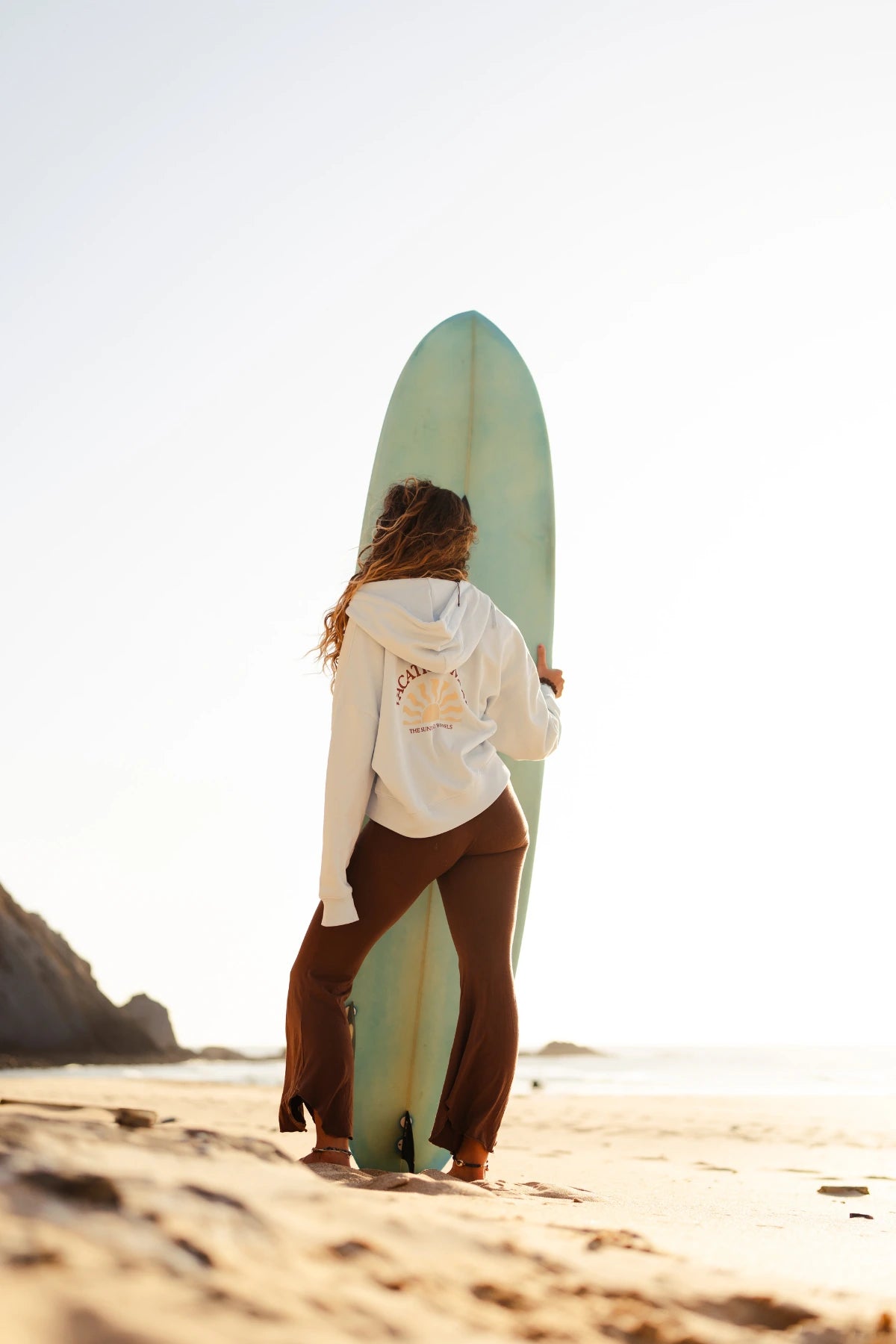 Damen Zip-Hoodie Iconic Women Pacific Chill in Cremeweiß mit großem Rückenprint, getragen am Strand neben einem Surfboard bei Sonnenuntergang.