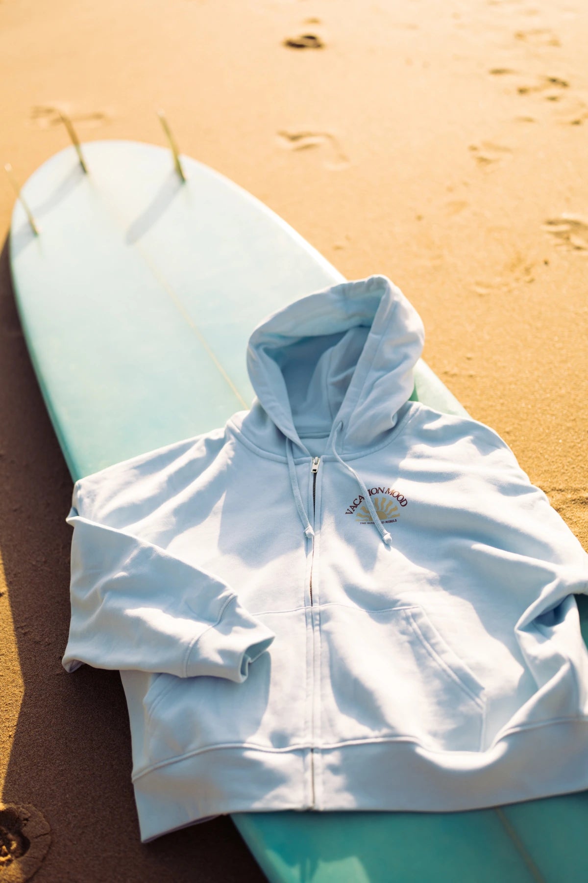 Iconic Women Zip-Hoodie Pacific Chill als Produktaufnahme auf einem Surfboard am Strand mit natürlichem Sonnenlicht.