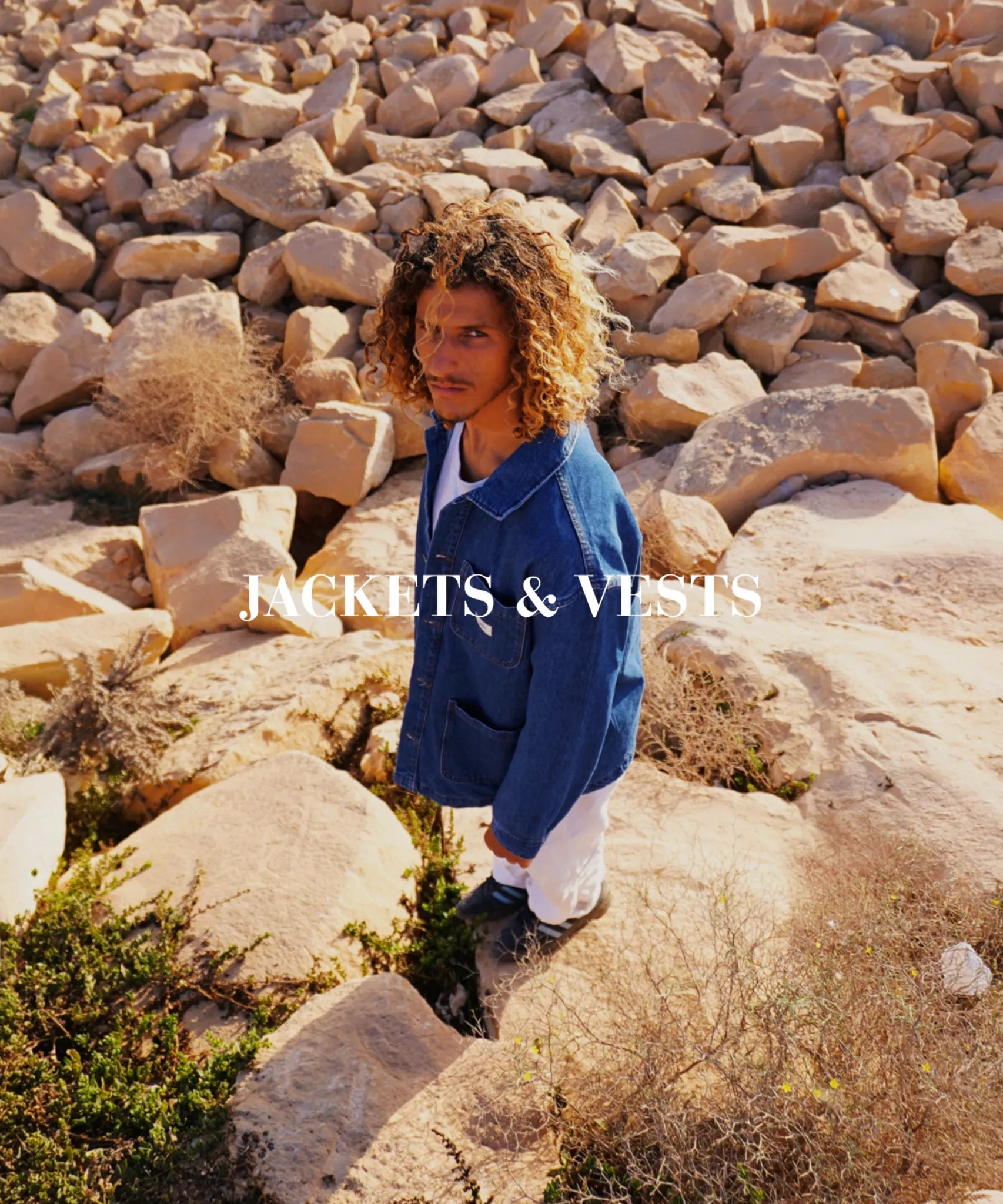 Person mit lockigen Haaren trägt blaue Jacke zwischen Felsen, warme Sonnenfarben, natürlicher Outdoor Look, Jackets und Vests Kategorie