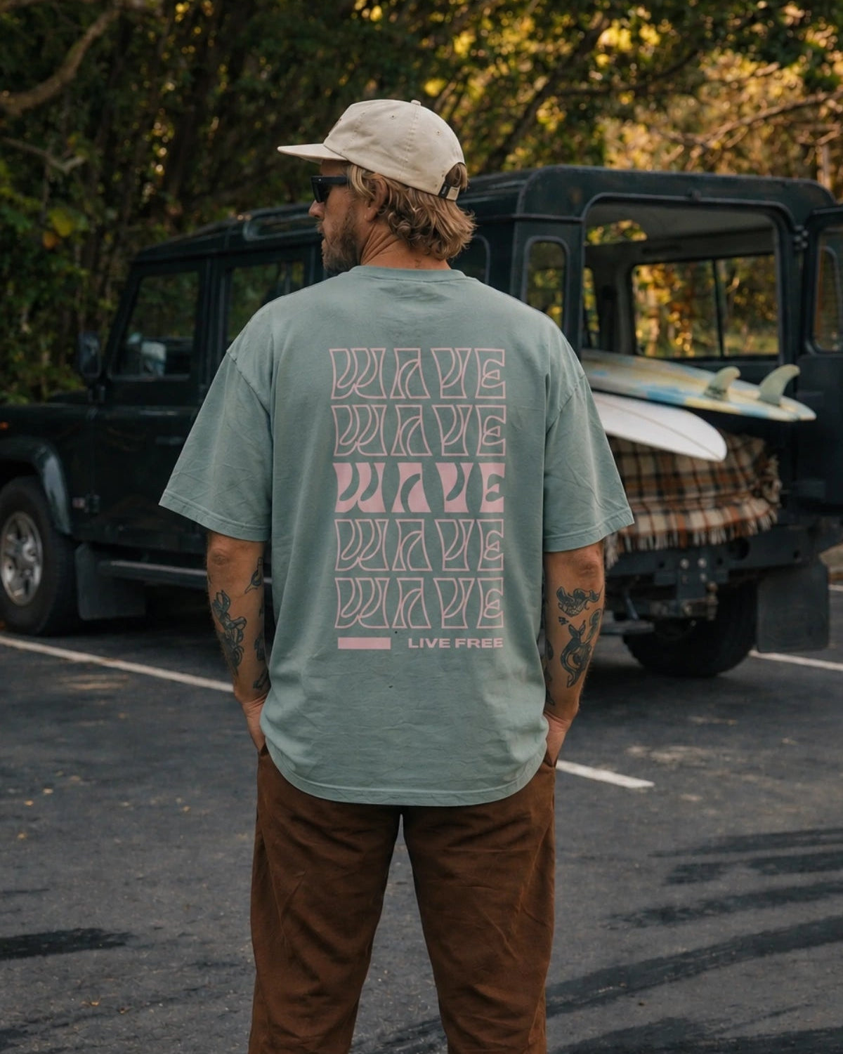 Mann von hinten im grünen Live Free Oversized T-Shirt Aloe mit rosa Wave Backprint vor Geländewagen und Surfboard – The Sunkissed Rebels