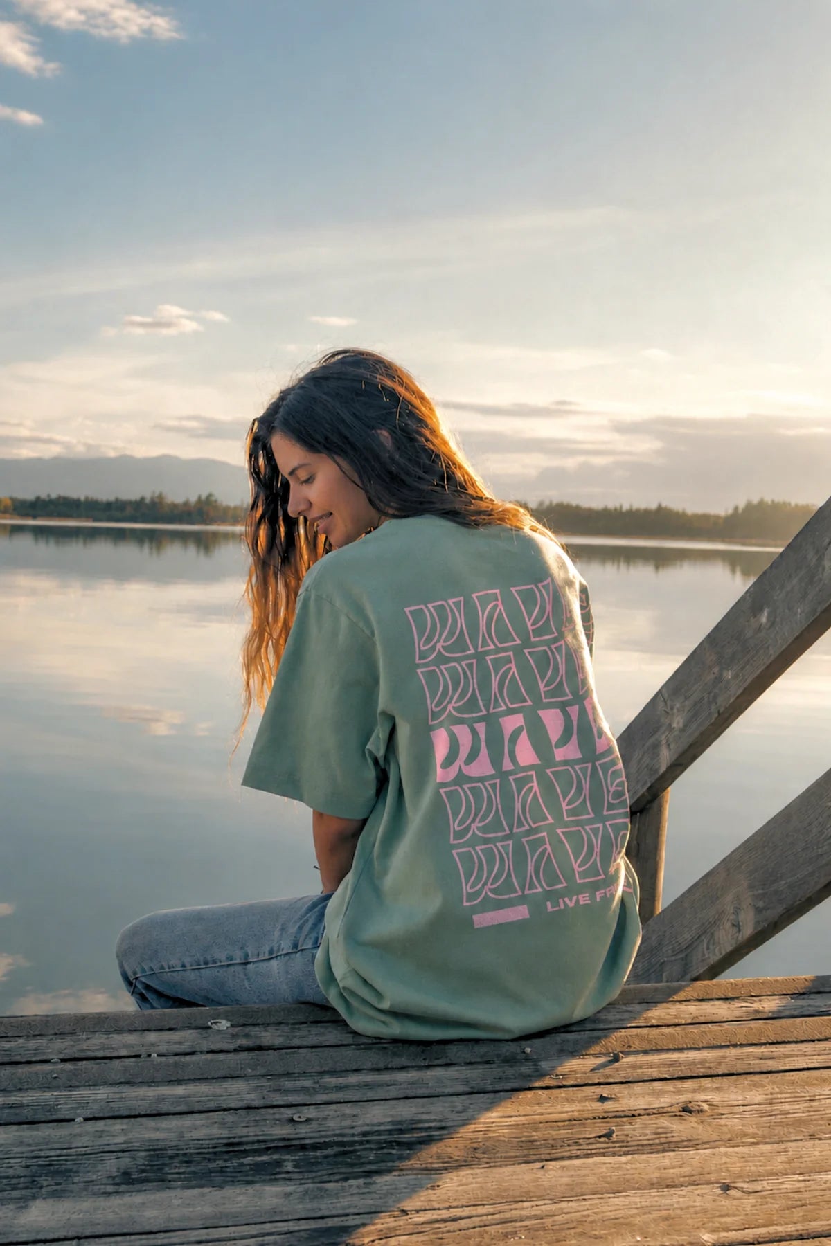 Rückenansicht des hellgrünen Live Free Oversized Shirts mit typografischem Wave-Print in Rosa auf einem Steg am Wasser.