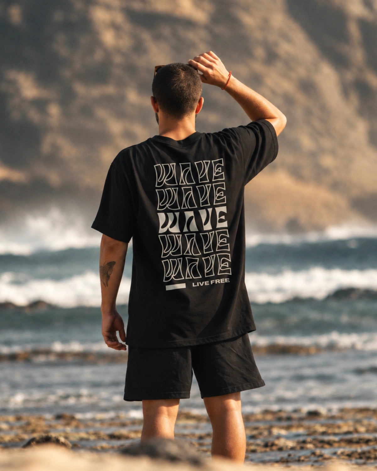 Mann trägt das schwarze Live Free Oversized T-Shirt von The Sunkissed Rebels mit großem Backprint am Strand