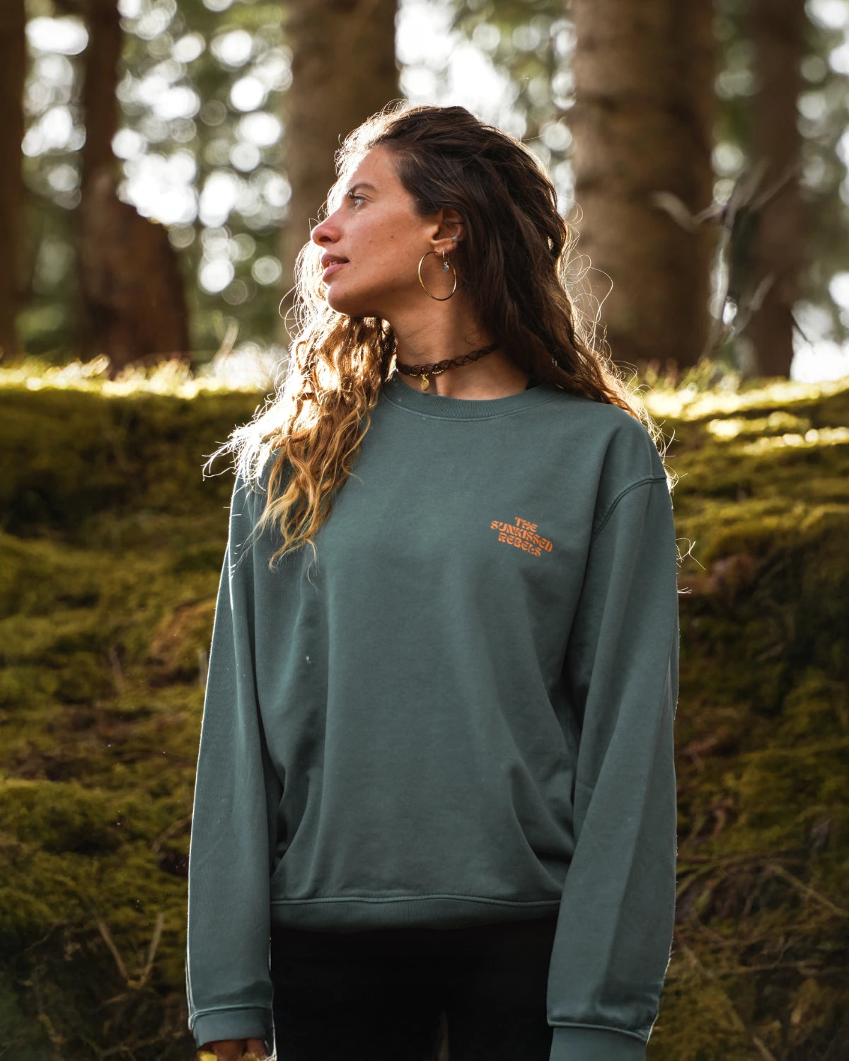 Frau trägt den Live Free Vintage Sweater in Palm Green mit kleinem orangem The Sunkissed Rebels Brustprint im Wald