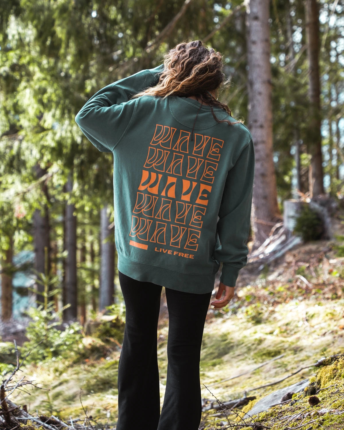 Frau trägt den Live Free Vintage Sweater in Palm Green von The Sunkissed Rebels mit großem orangem Rückenprint im Wald