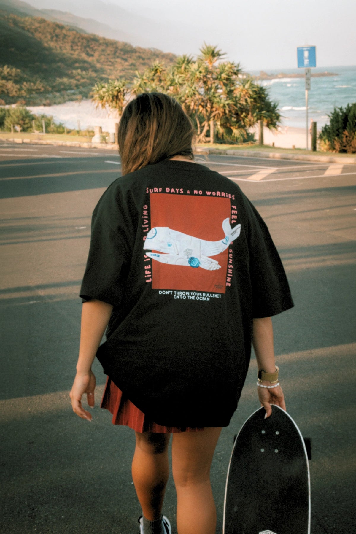 Frau trägt das No Bullshit Whale Boxy Oversized T-Shirt in Black von The Sunkissed Rebels mit großem Whale Backprint und Skateboard.