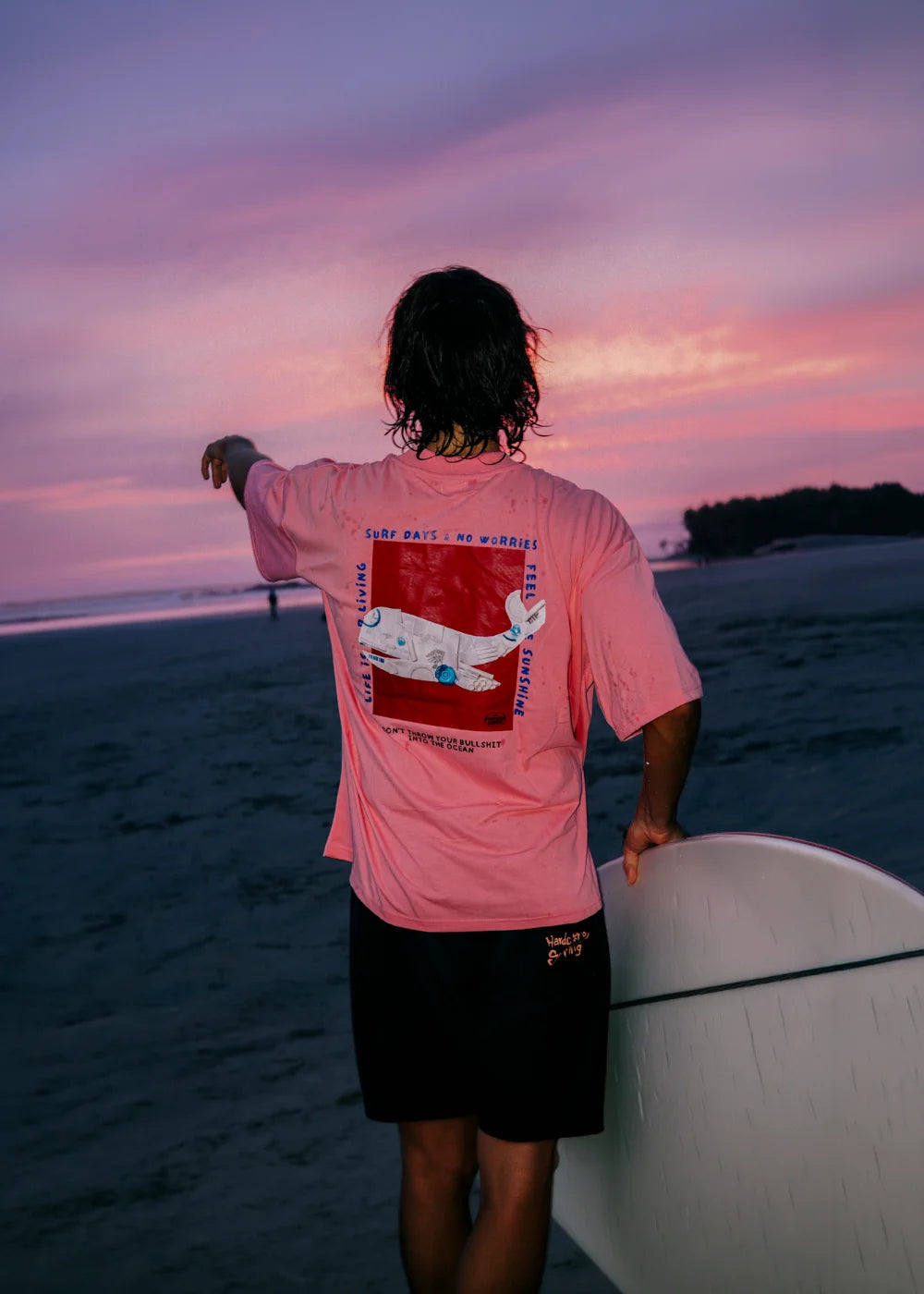 Surfer trägt das pinke Surf Days Oversized T-Shirt mit großem Rückenprint von The Sunkissed Rebels bei Sonnenuntergang am Strand mit Surfboard.