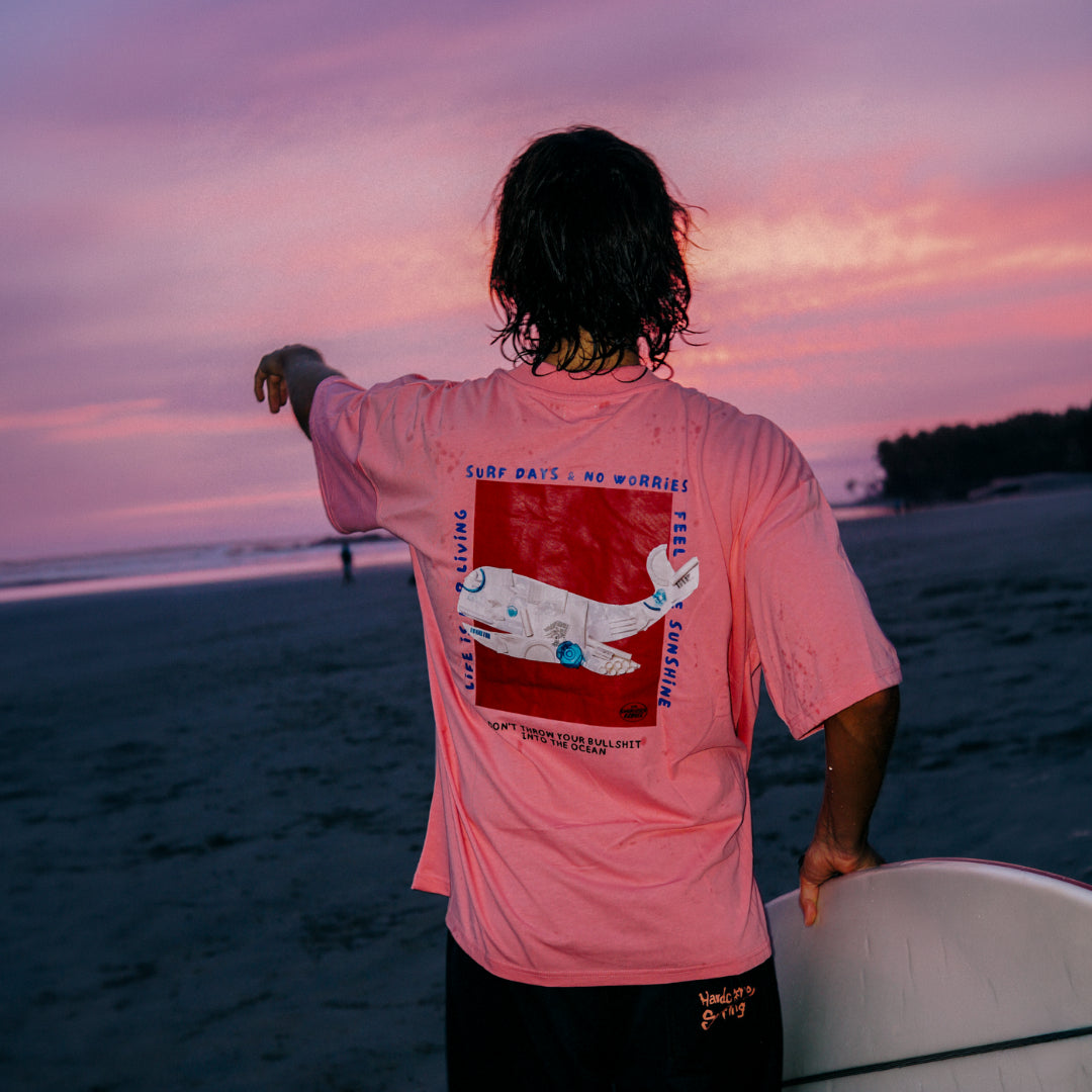 Rückansicht eines Surfers im pinken No Bullshit Oversized T-Shirt Pink – The Sunkissed Rebels mit Surfboard bei Sonnenuntergang.