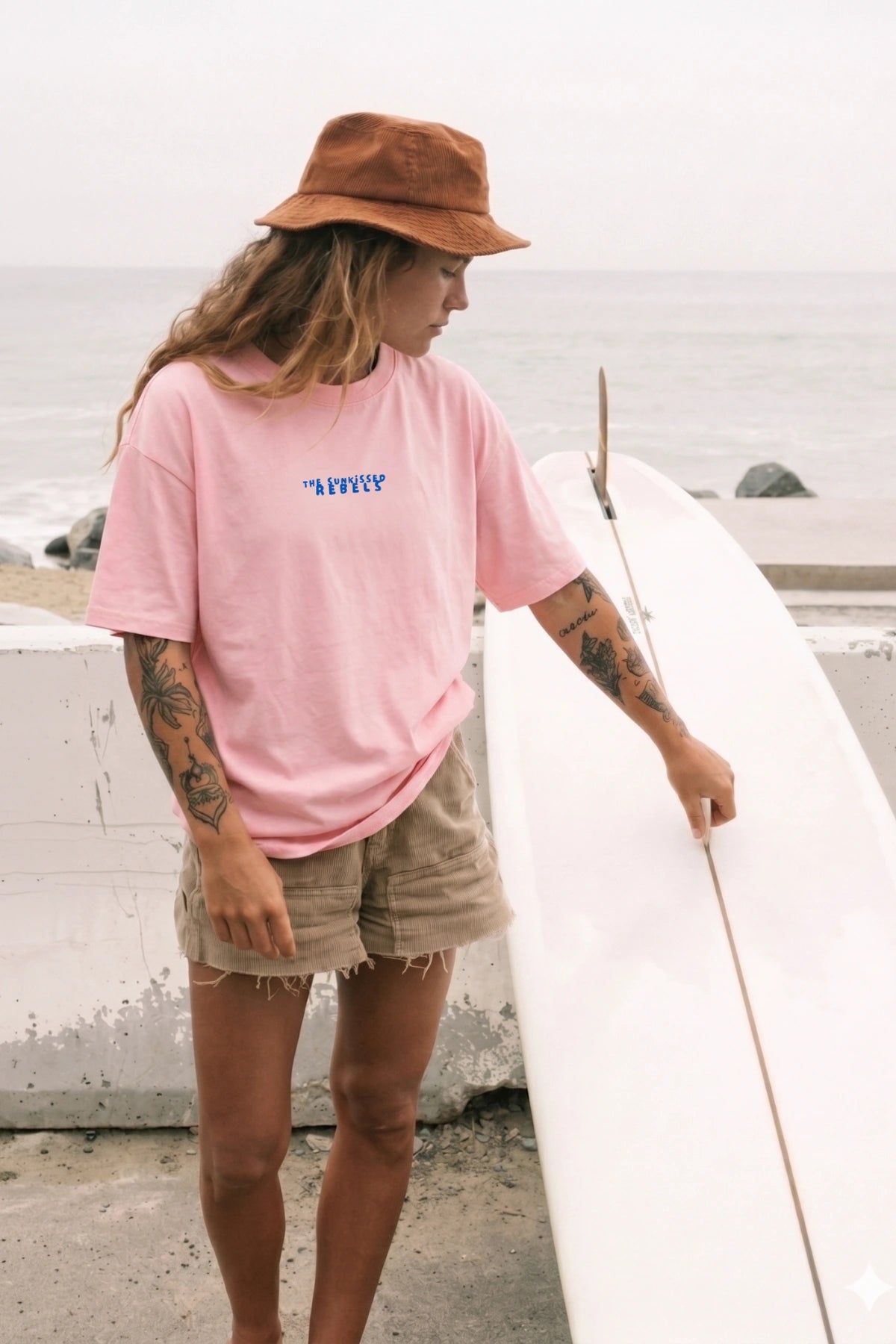 Model trägt pinkes Oversized Surf T Shirt am Strand neben weißem Surfboard im California Beach Style