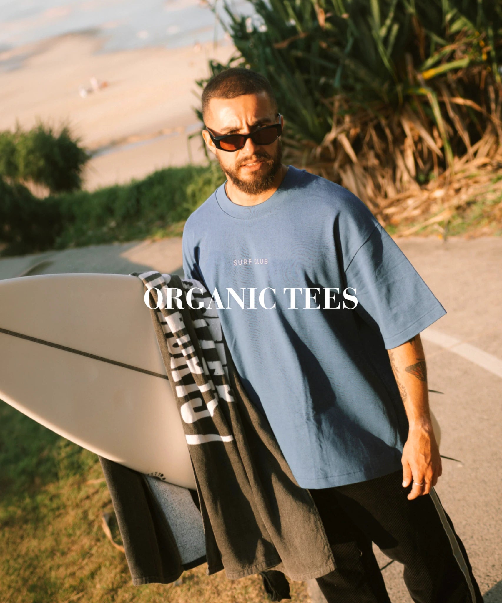 Mann mit Surfboard trägt blaues T-Shirt am Strand, Sonnenlicht und entspannter Surf Lifestyle, Organic Tees Kategorie