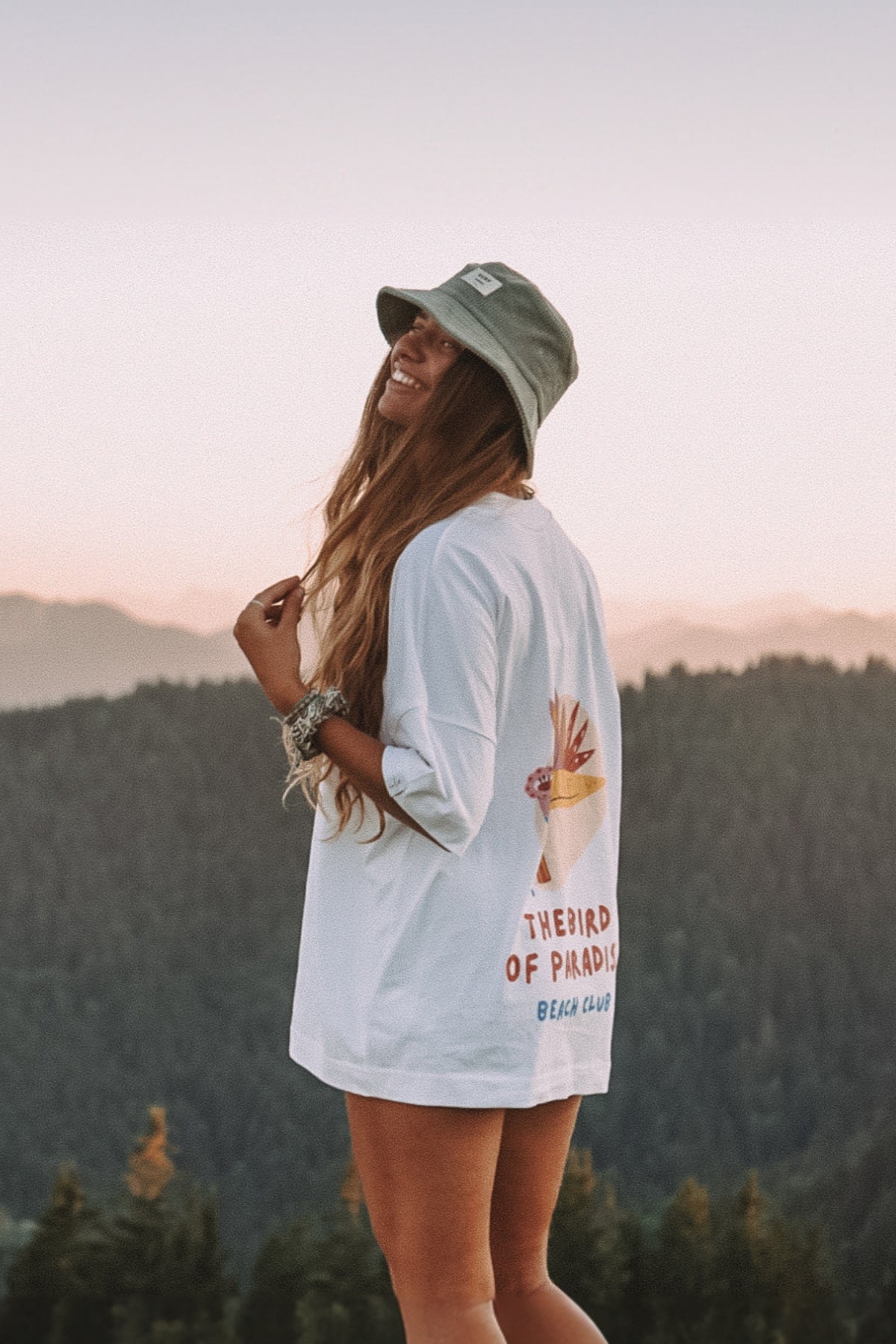 Frau steht auf Berg, lacht und trägt dabei das oversized T-Shirt Bird of Paradise in weiß von The Sunkissed Rebels